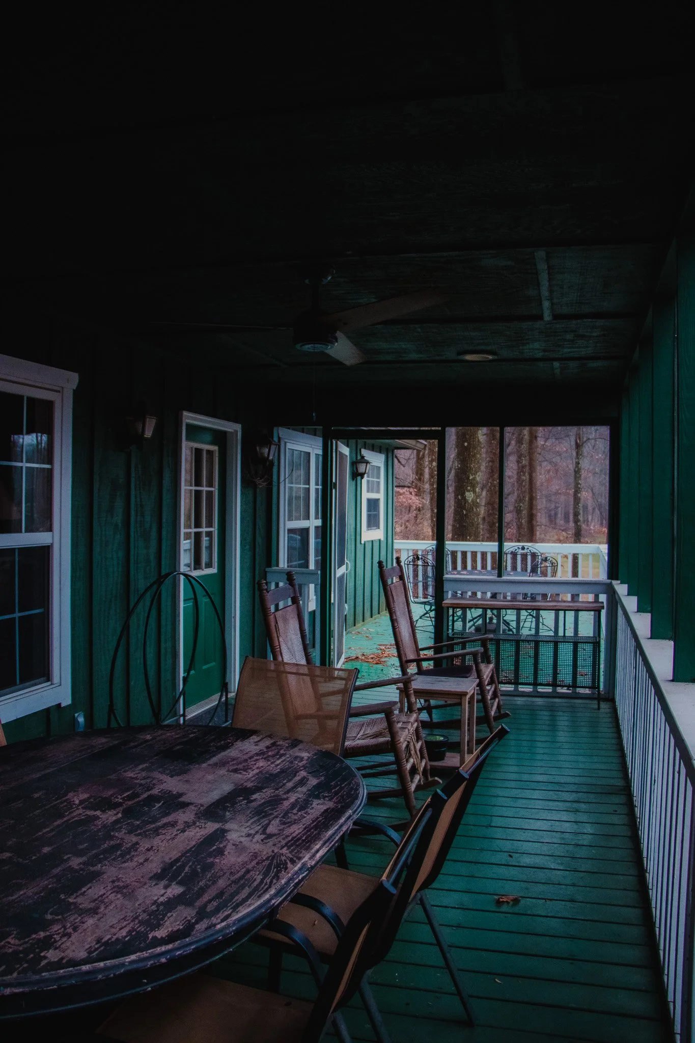 An outdoor porch with a green wooden floor and a table surrounded by several chairs, some of which are wooden rocking chairs. The porch has a roof, ceiling fan, and white railing, with trees in the background.