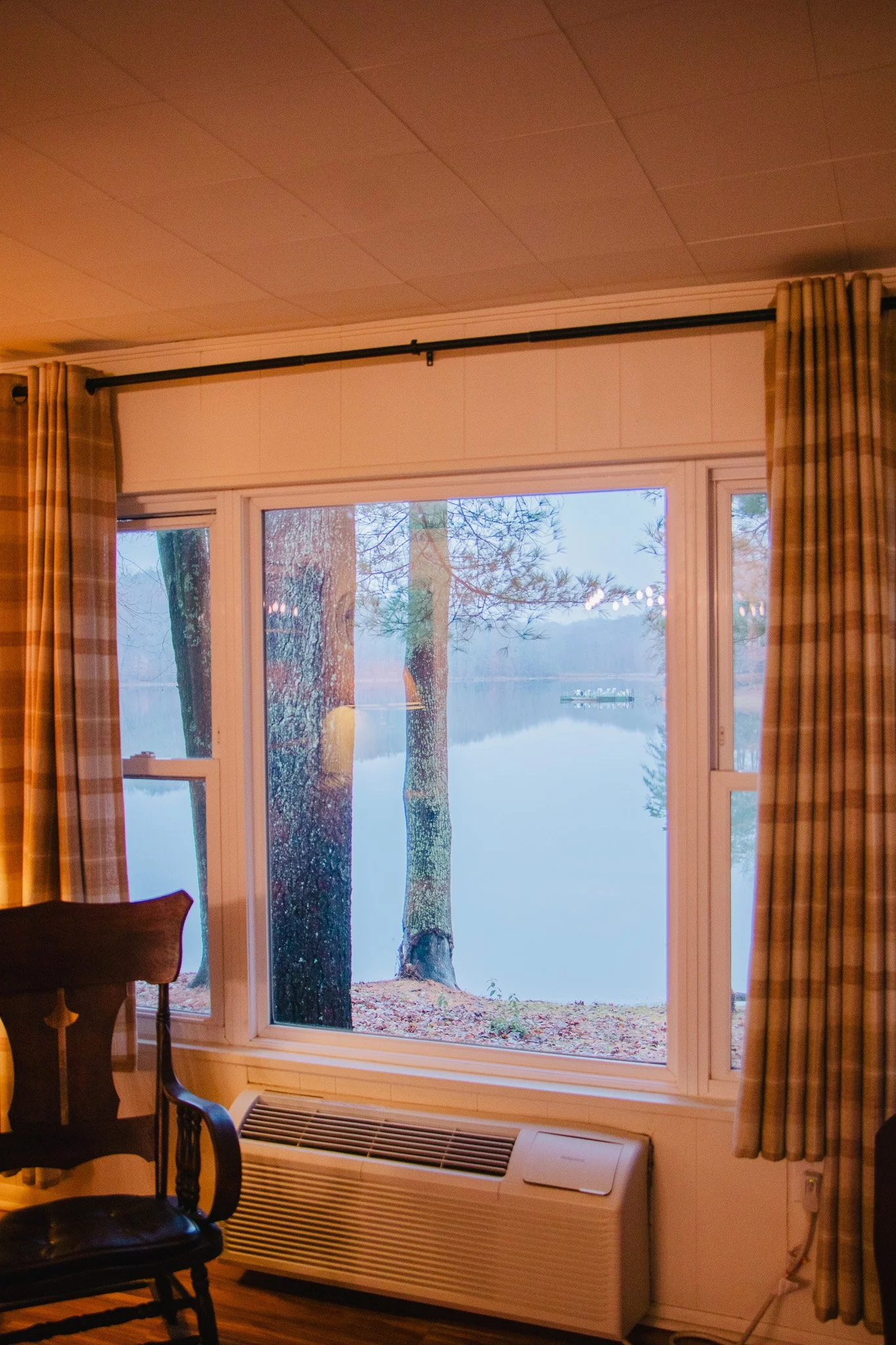 View of a lake with trees outside a large window in a room, with wooden flooring, curtains, and a chair inside.
