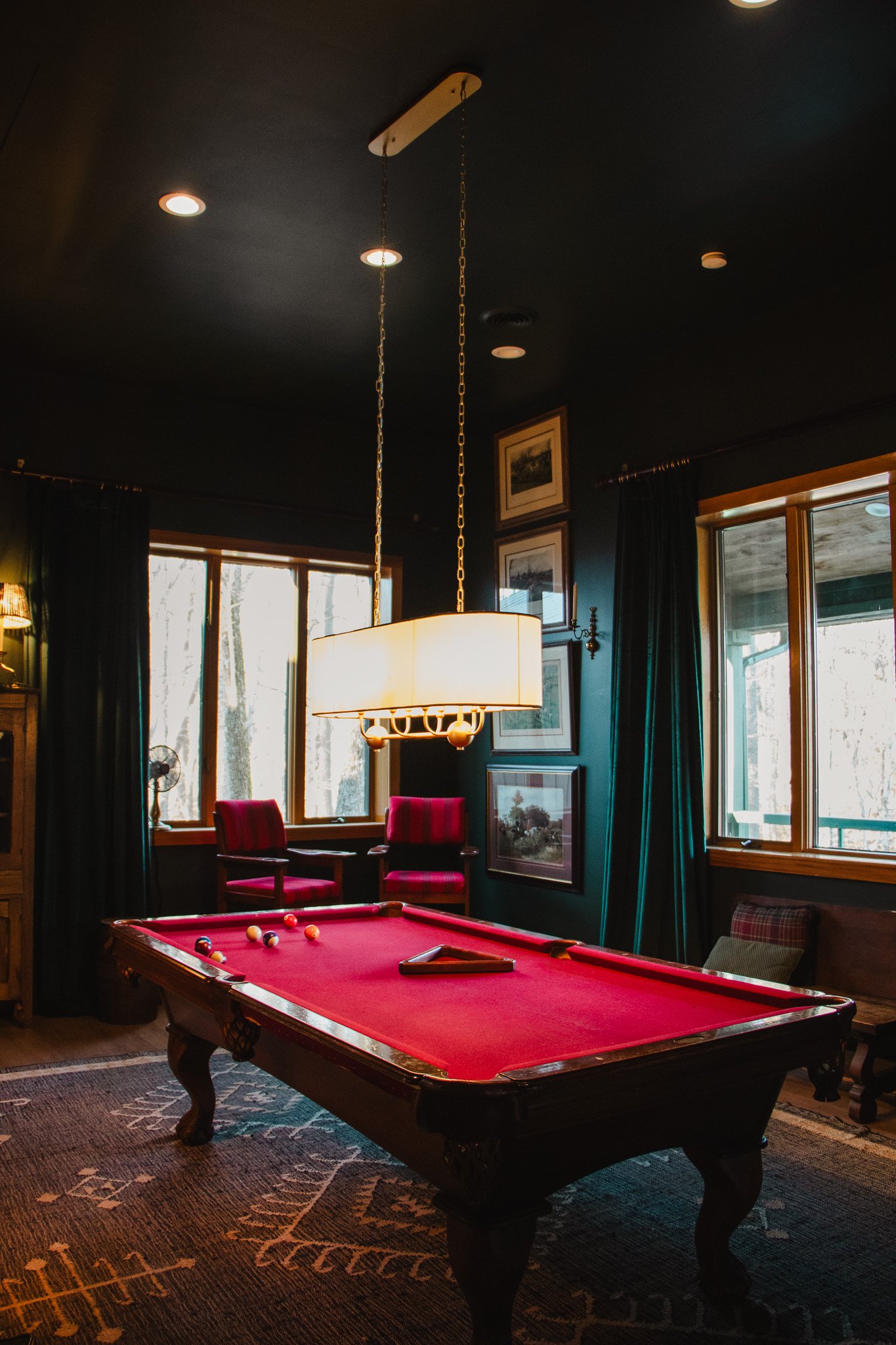 A cozy room with a red pool table, two red chairs by the window, dark curtains, framed pictures on the wall, and a hanging light fixture. Sunlight streams through large windows, revealing a wooded outdoor view.