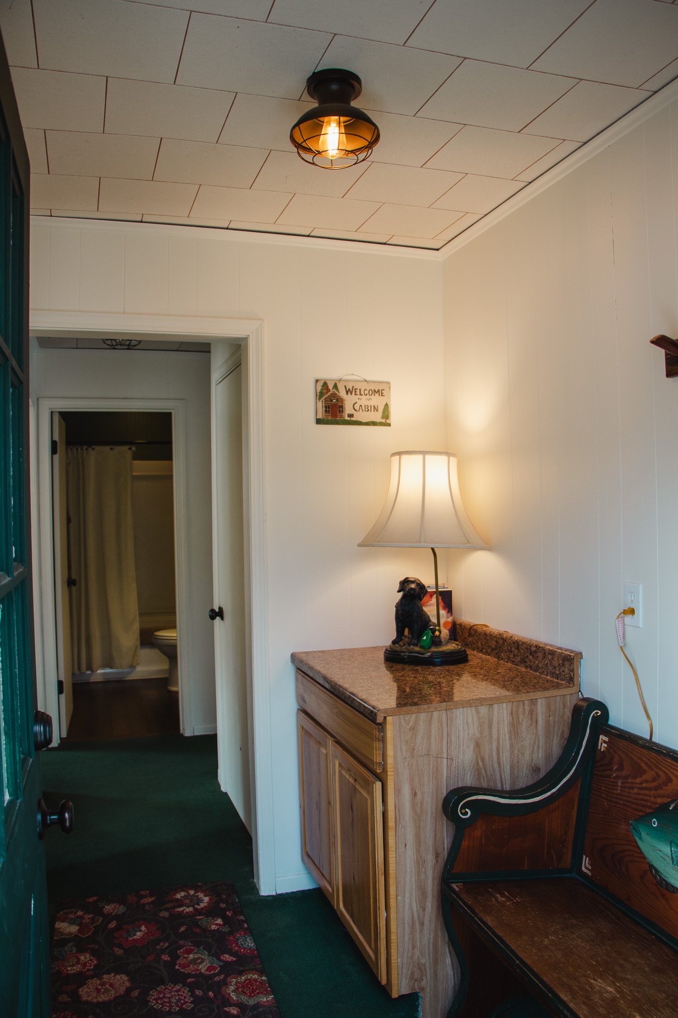 Interior of a cozy cabin bedroom with a small wooden cabinet, a table lamp with a beige shade, and a decorative dog figurine on the cabinet. Part of a wooden bench is visible on the right. In the background, there is a doorway leading to a bathroom w