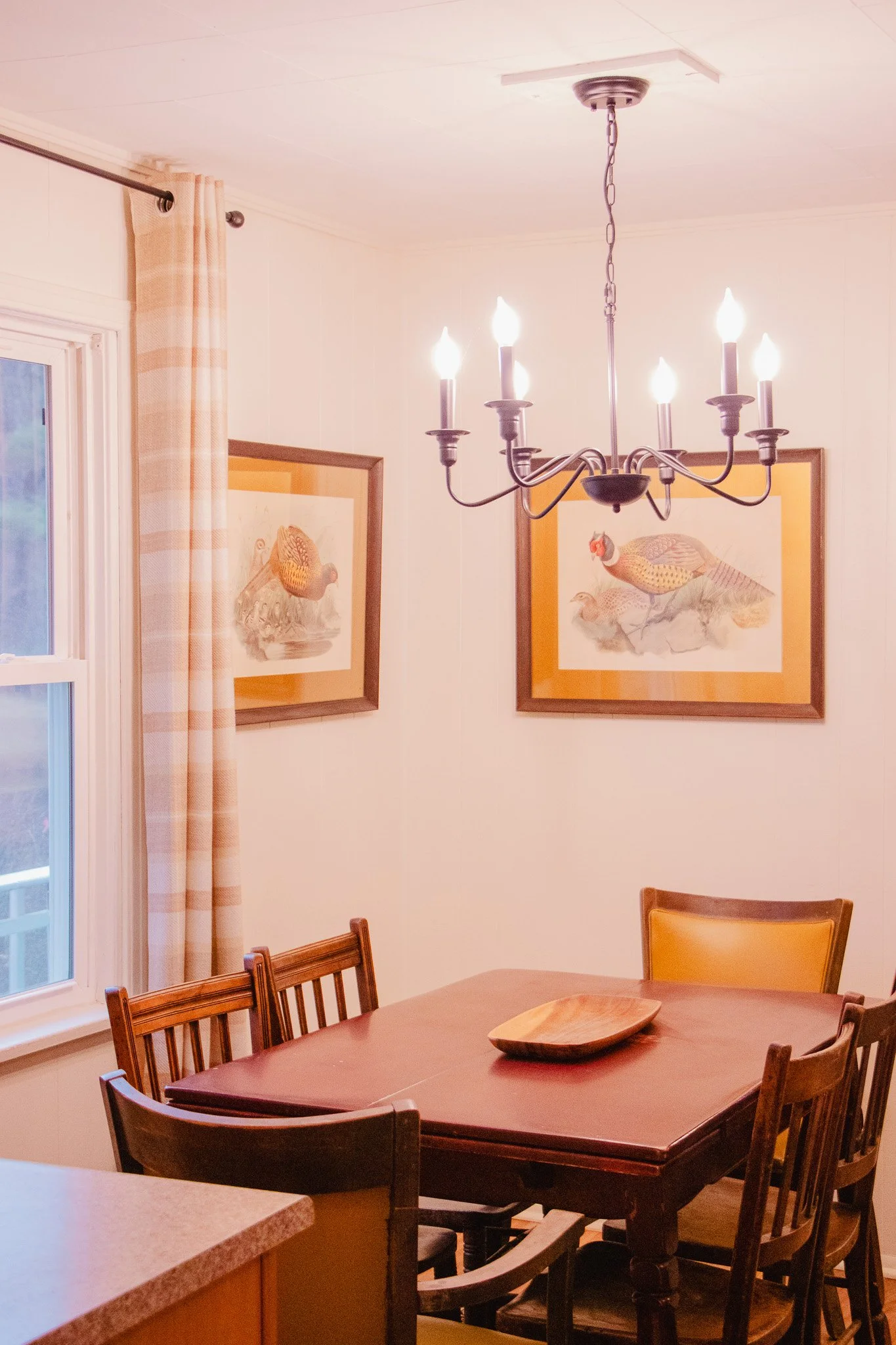 A dining room with a wooden table and chairs, a hanging chandelier, framed artwork on the walls depicting birds, a window with curtains, and a wooden bowl on the table.