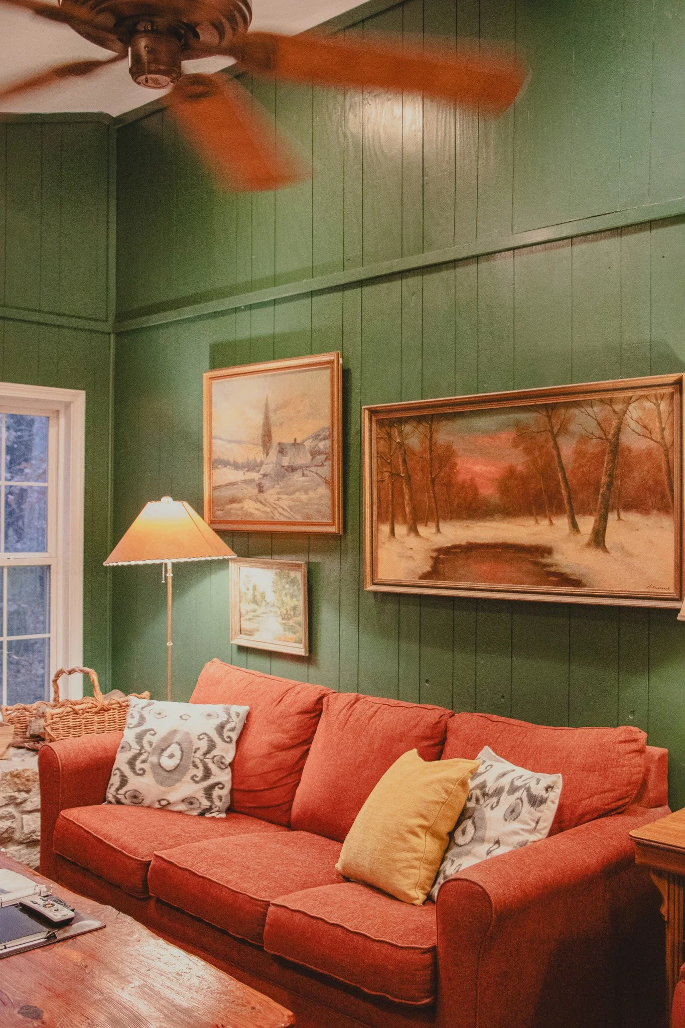 Living room corner with green wooden paneled walls, a red sofa with yellow and patterned cushions, a floor lamp with a beige shade, a wooden coffee table, and three landscape paintings hanging on the wall, one of which depicts a snowy scene with tree