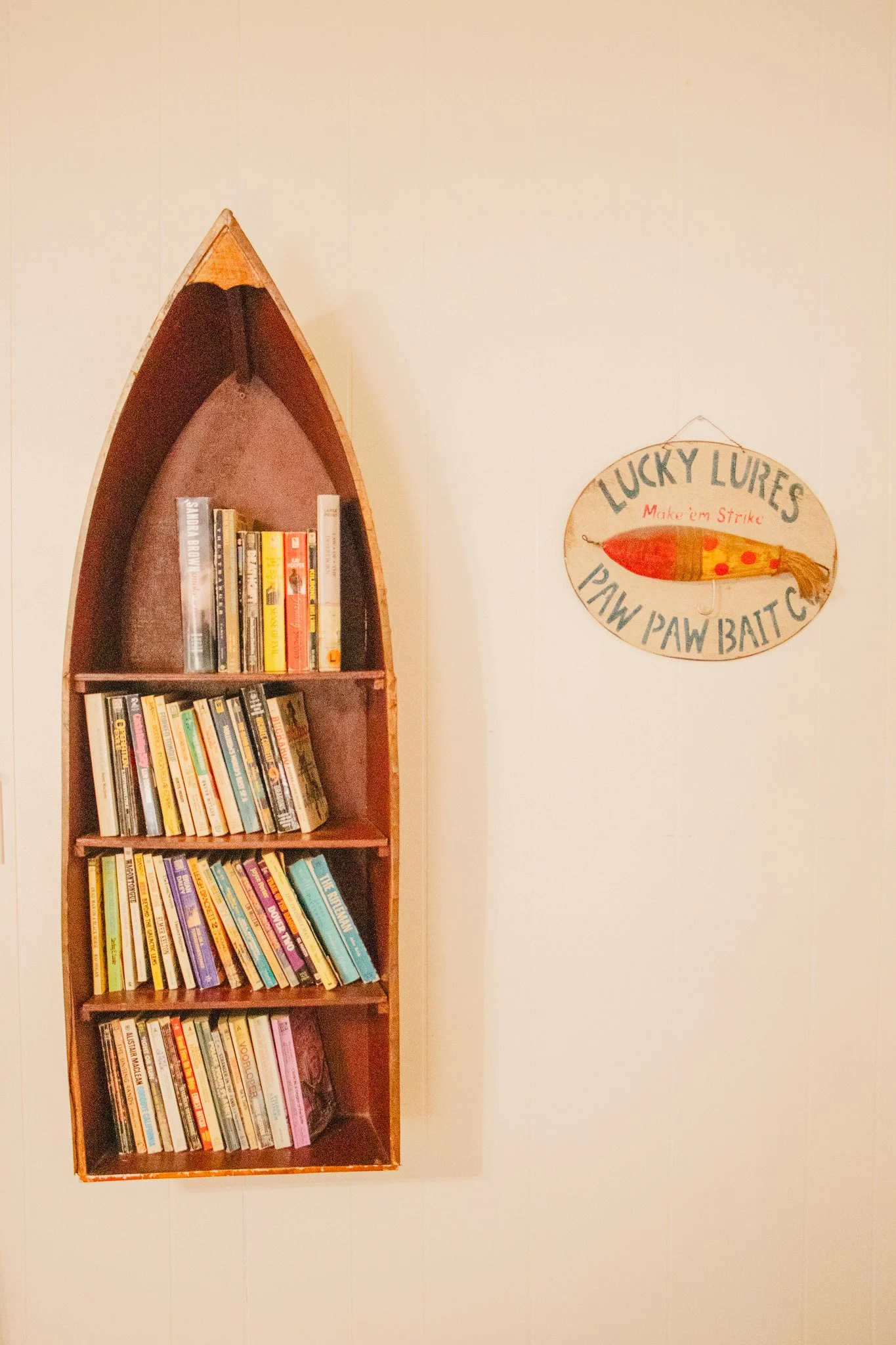 A bookshelf shaped like a boat filled with books, mounted on a wall. To the right, a sign reads 'Lucky Lures Make 'em Strike Paw Paw Bait' with an illustration of a fishing lure.