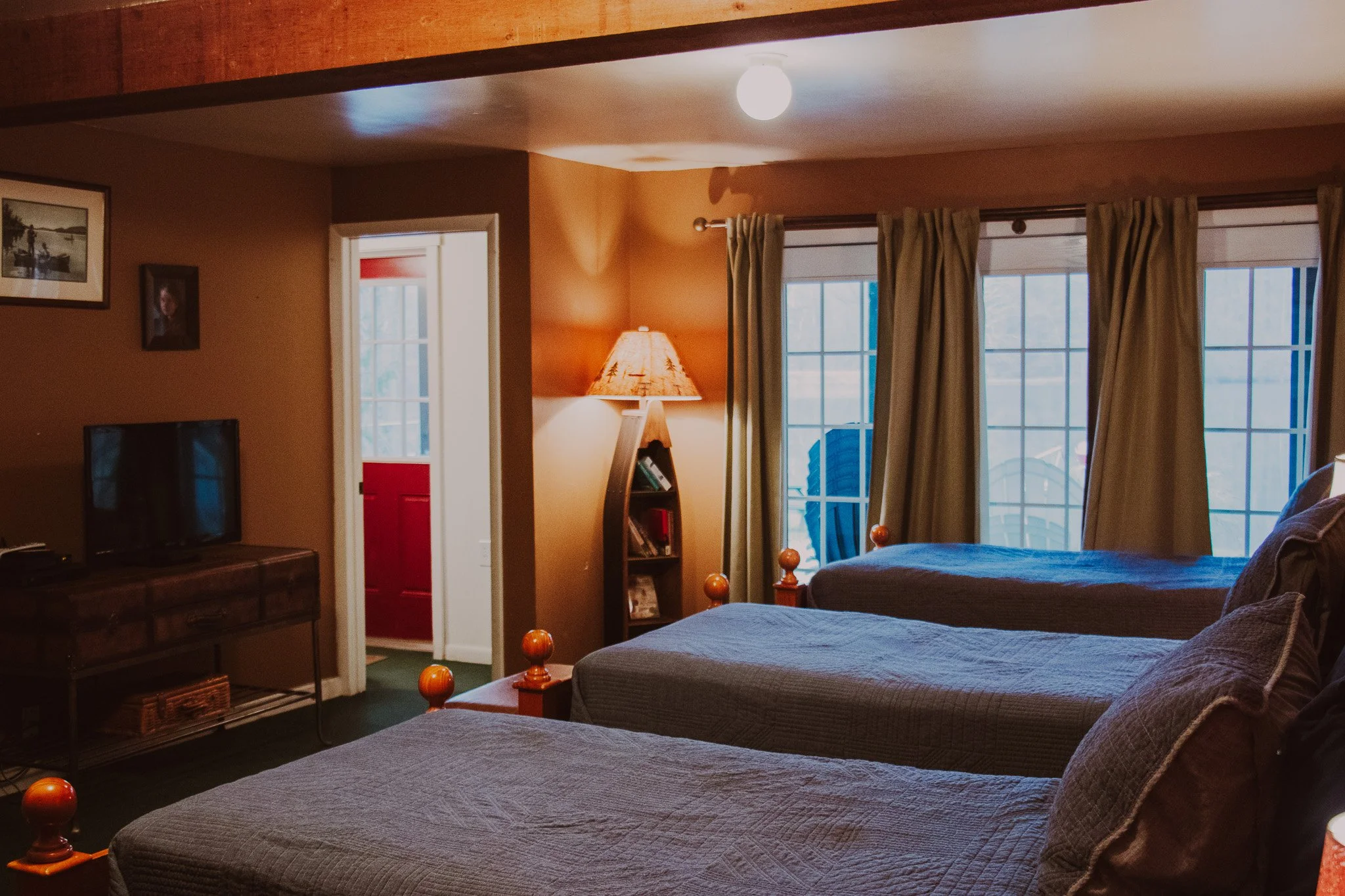 A cozy bedroom with three beds, a sliding glass door with curtains leading to a deck, a standing lamp, a TV on a wooden stand, and framed pictures on the wall.