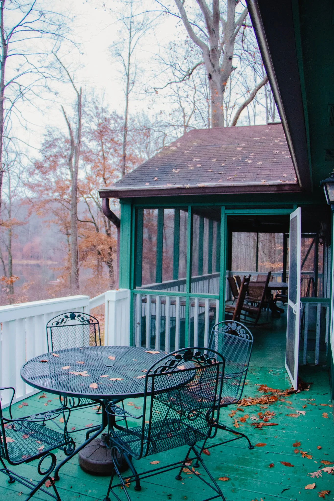 Image of a porch with a round metal table and four matching chairs, all covered with fallen leaves. In the background, there is a house with a screened-in porch area and a doorway open to the outside. The house is painted green with white trim, and t