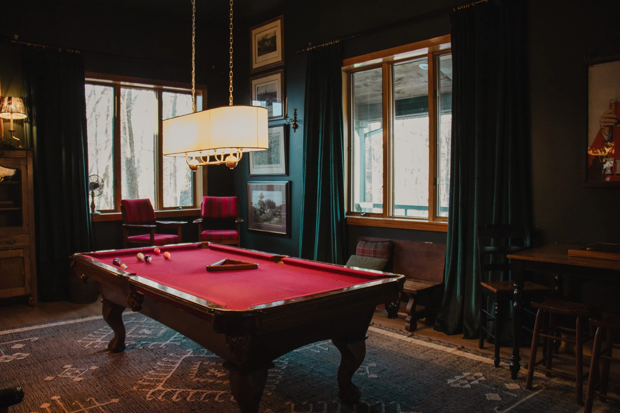 A cozy game room with a red pool table, two red armchairs, and dark green curtains by windows showing trees outside. There is a large hanging light fixture above the pool table and some artwork on the walls.