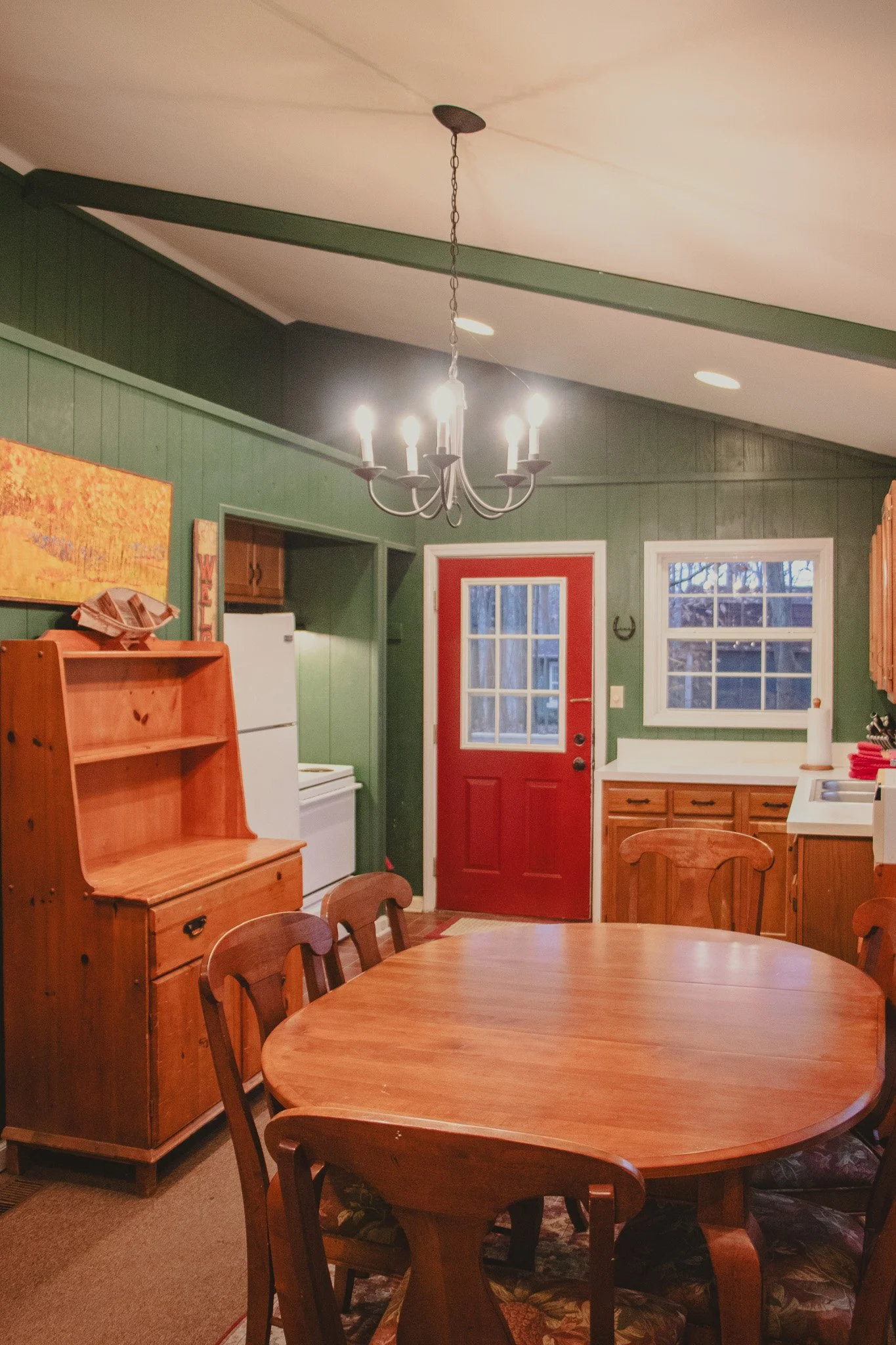A cozy kitchen and dining area with green paneled walls, a red door, and two windows. Wooden furniture, including a round dining table with chairs, a hutch, and cabinets. Chandelier hanging from the ceiling and a white refrigerator.