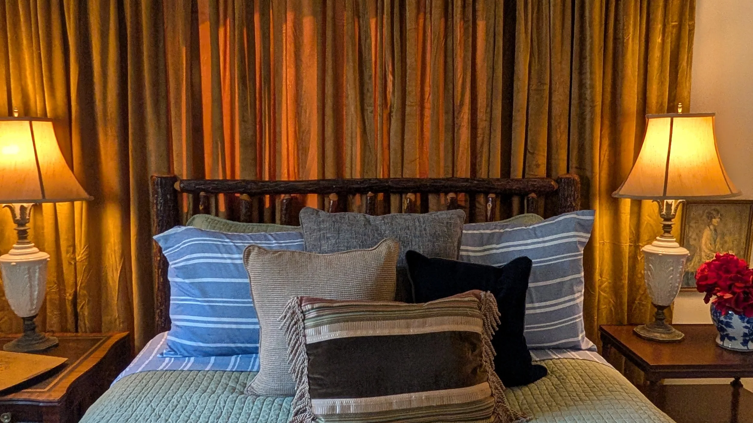 A cozy bedroom with a bed, two pillows, a wooden headboard, a bedside table, a lamp, white curtains, and windows showing an outdoor scene.