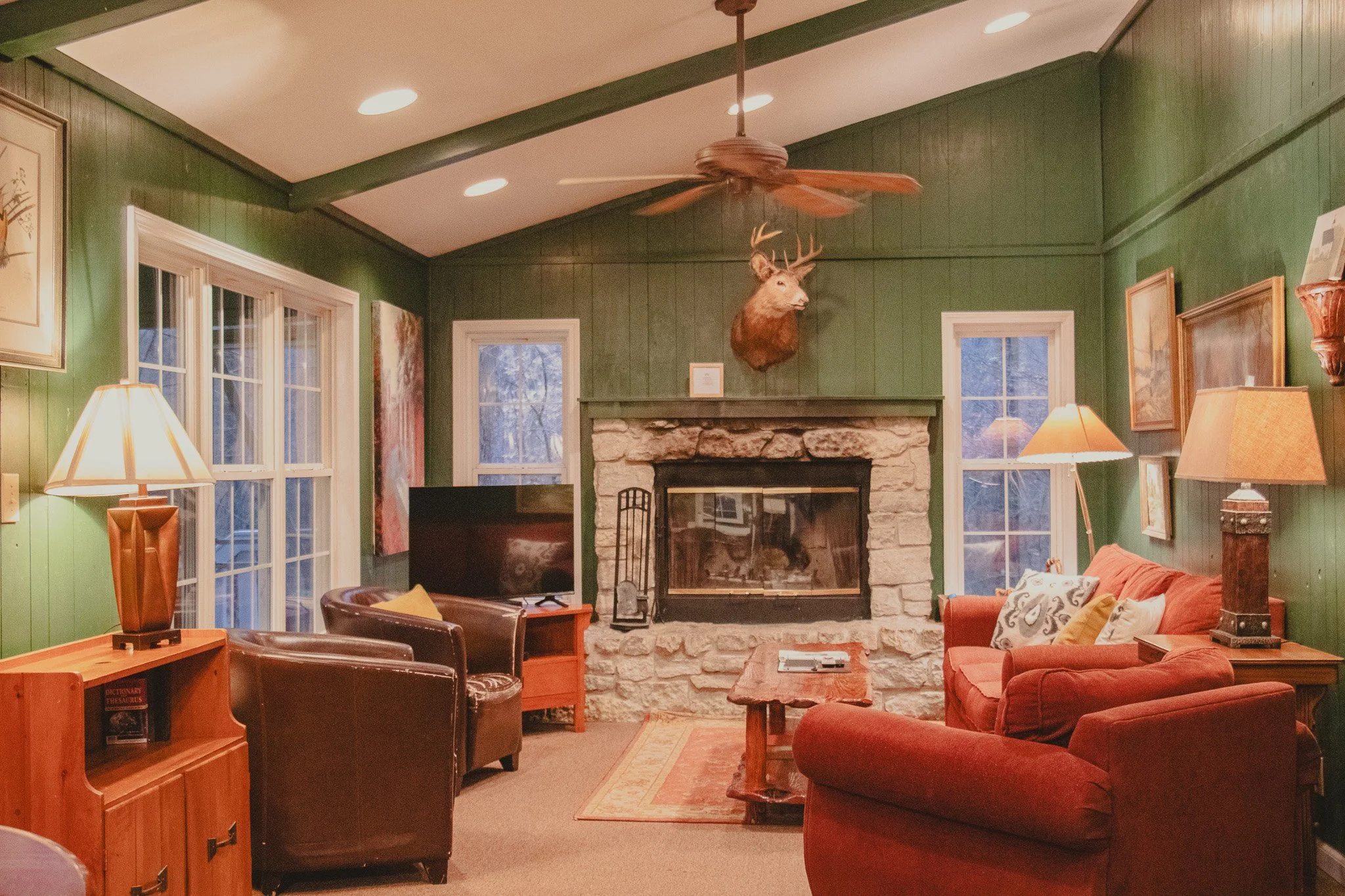 Cozy living room with green wooden walls, stone fireplace, mounted deer head, ceiling fan, and various chairs and lamps.
