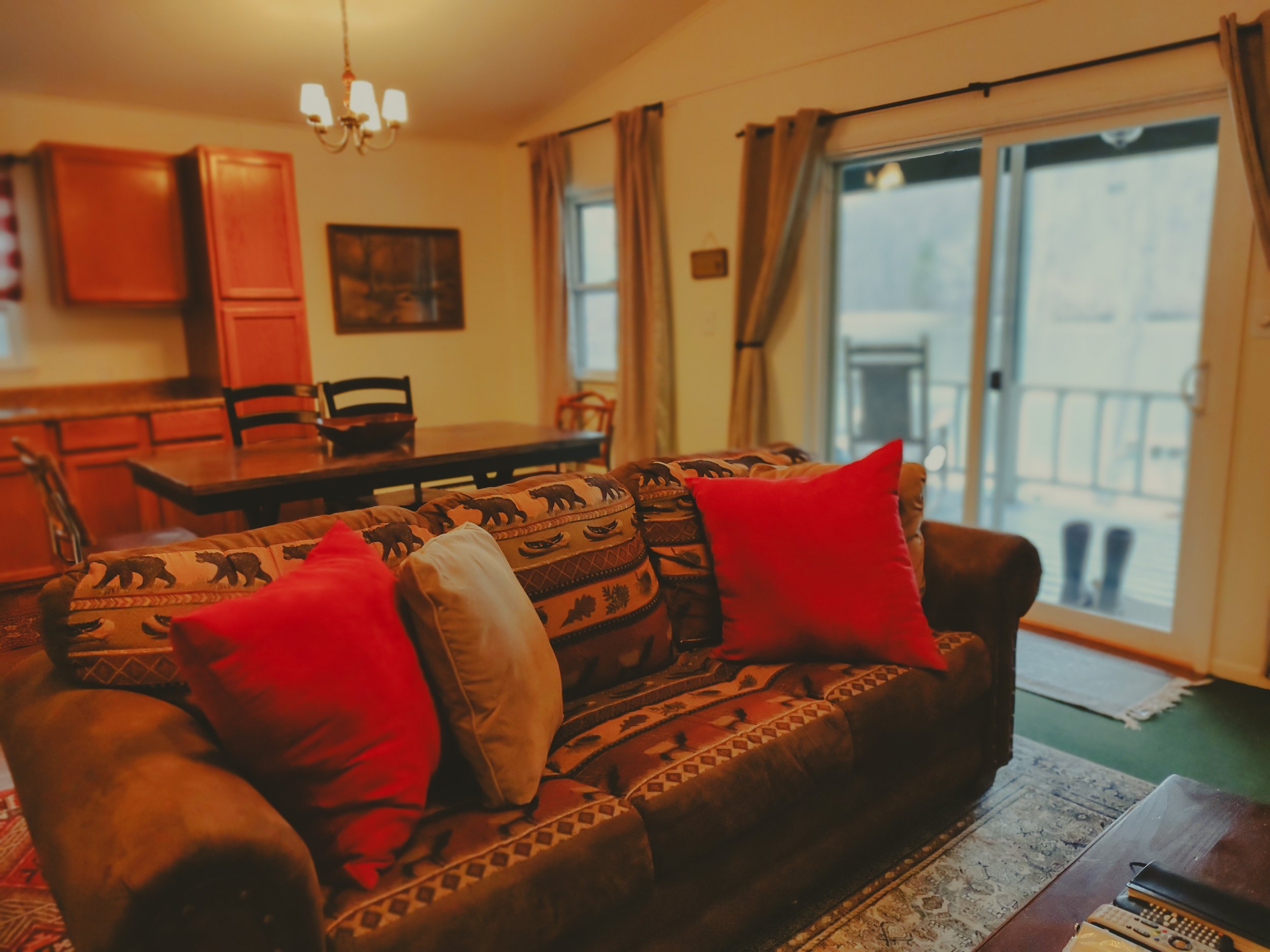 Cozy living room with patterned sofa and red and beige pillows, wooden dining table with chairs, sliding glass door leading to balcony, and warm lighting.