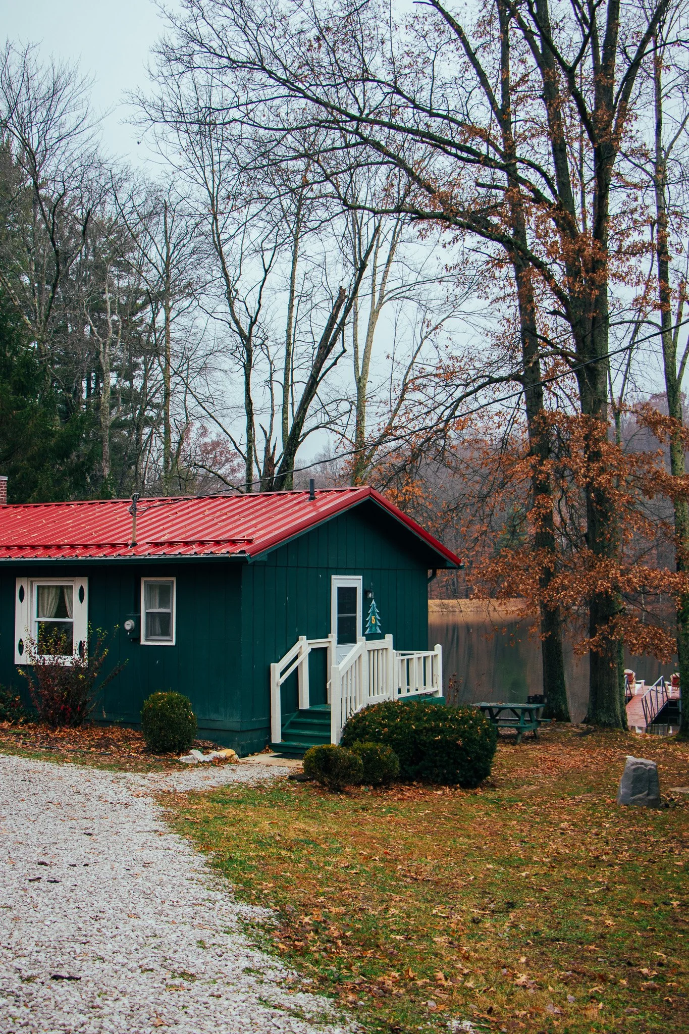 A small teal-colored house with a red metal roof, surrounded by trees with autumn leaves. There are bushes and a gravel path leading to a white door with a small porch. In the background, there's a lake and a dock.