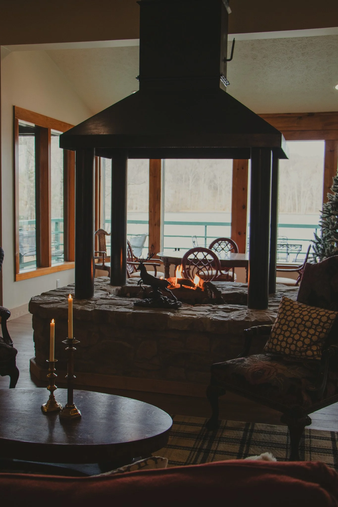 Living room with a stone fireplace, lit with a fire, surrounded by wooden furniture and decorated for the holidays, with large windows showing a view outside.