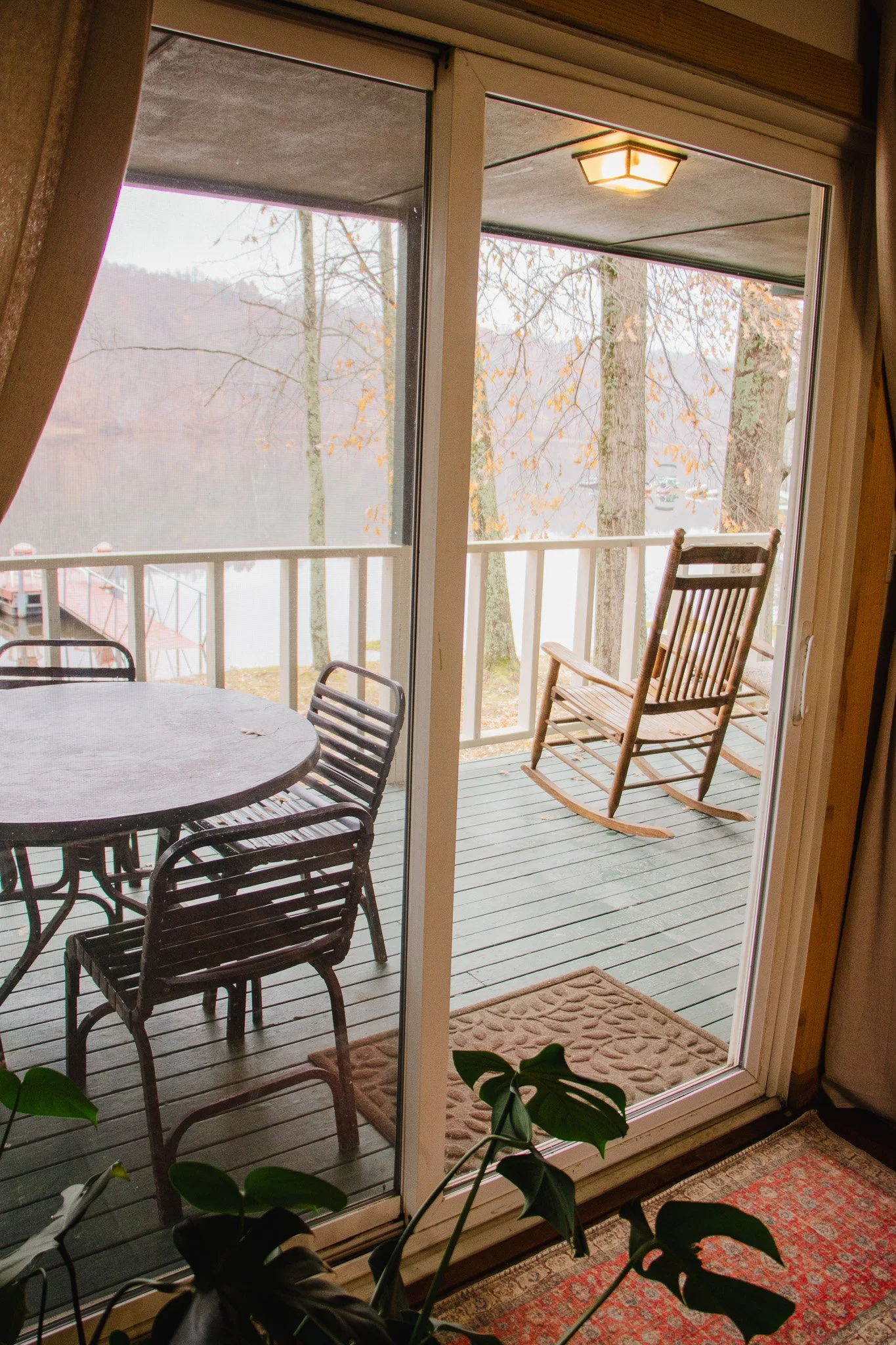 Glass sliding door leading to a balcony with wooden rocking chair, metal table, and chairs on a wooden deck overlooking a river and trees with autumn leaves.