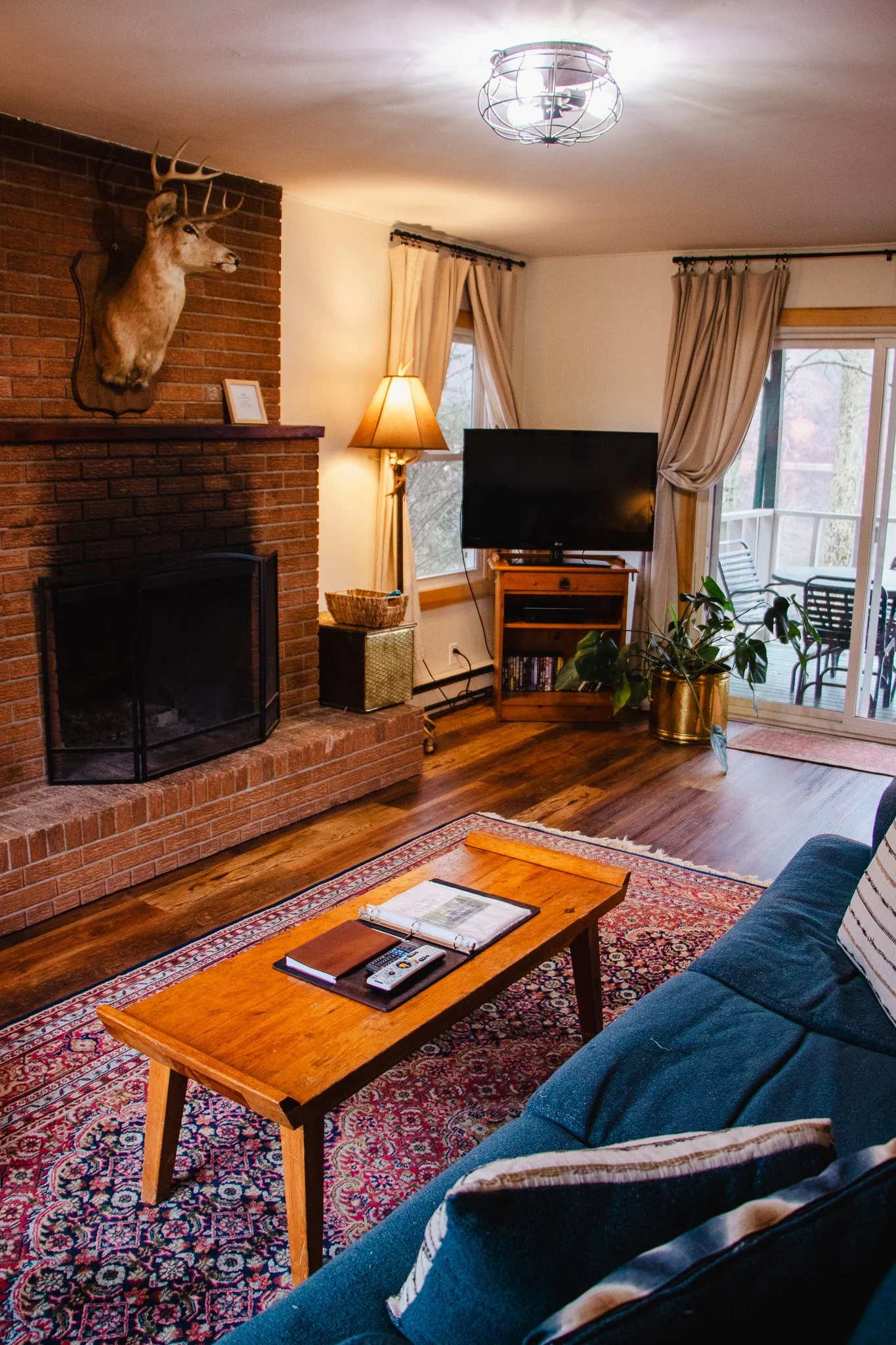 Living room with brick fireplace, mounted deer head, wooden coffee table, blue couch, patterned rug, flat-screen TV on wooden stand, large potted plant, sliding glass door with curtains leading to balcony.