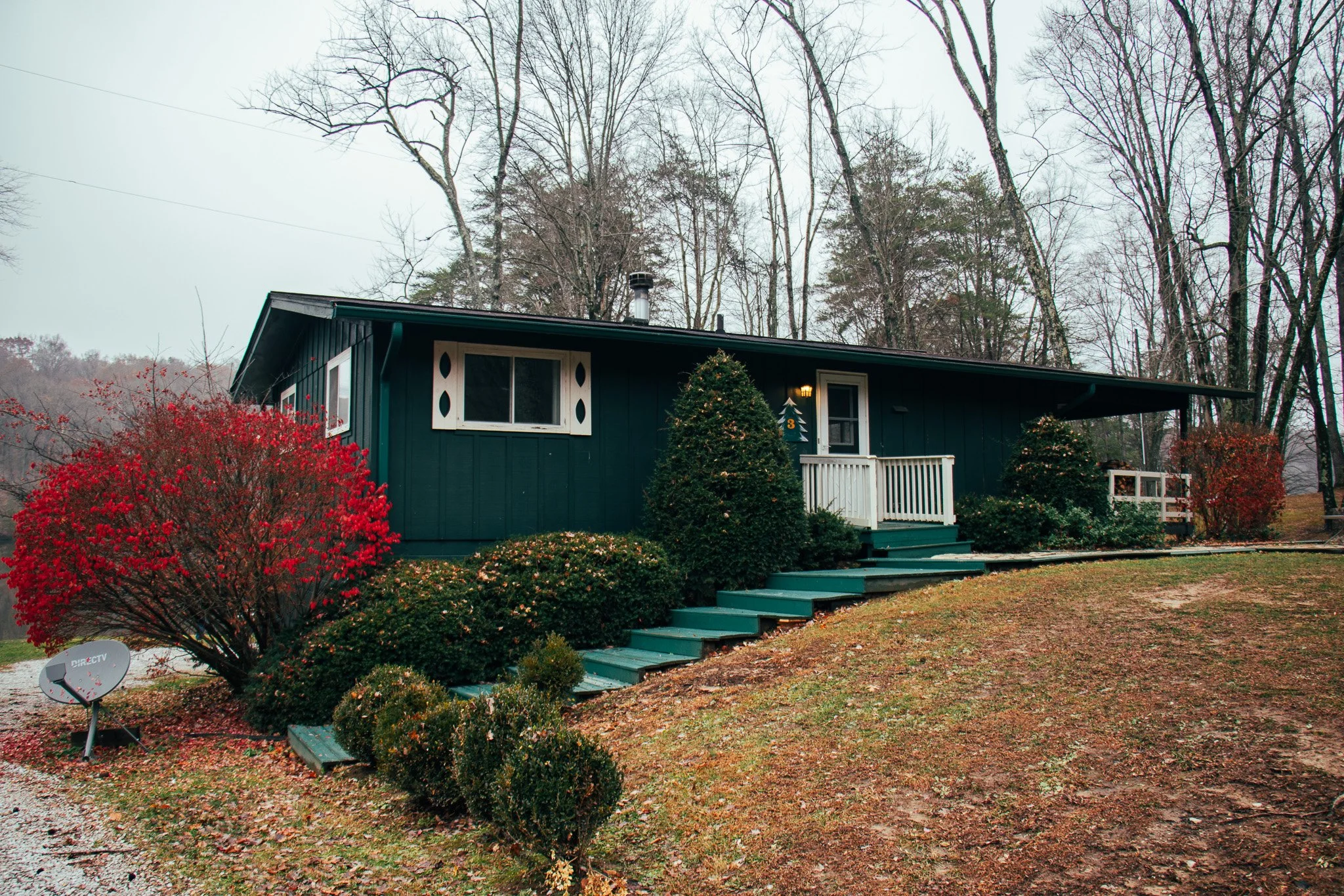 A house painted dark green with white trim, a small front porch, and steps leading up to the entrance, surrounded by leafless trees and bushes, with a satellite dish in the yard.