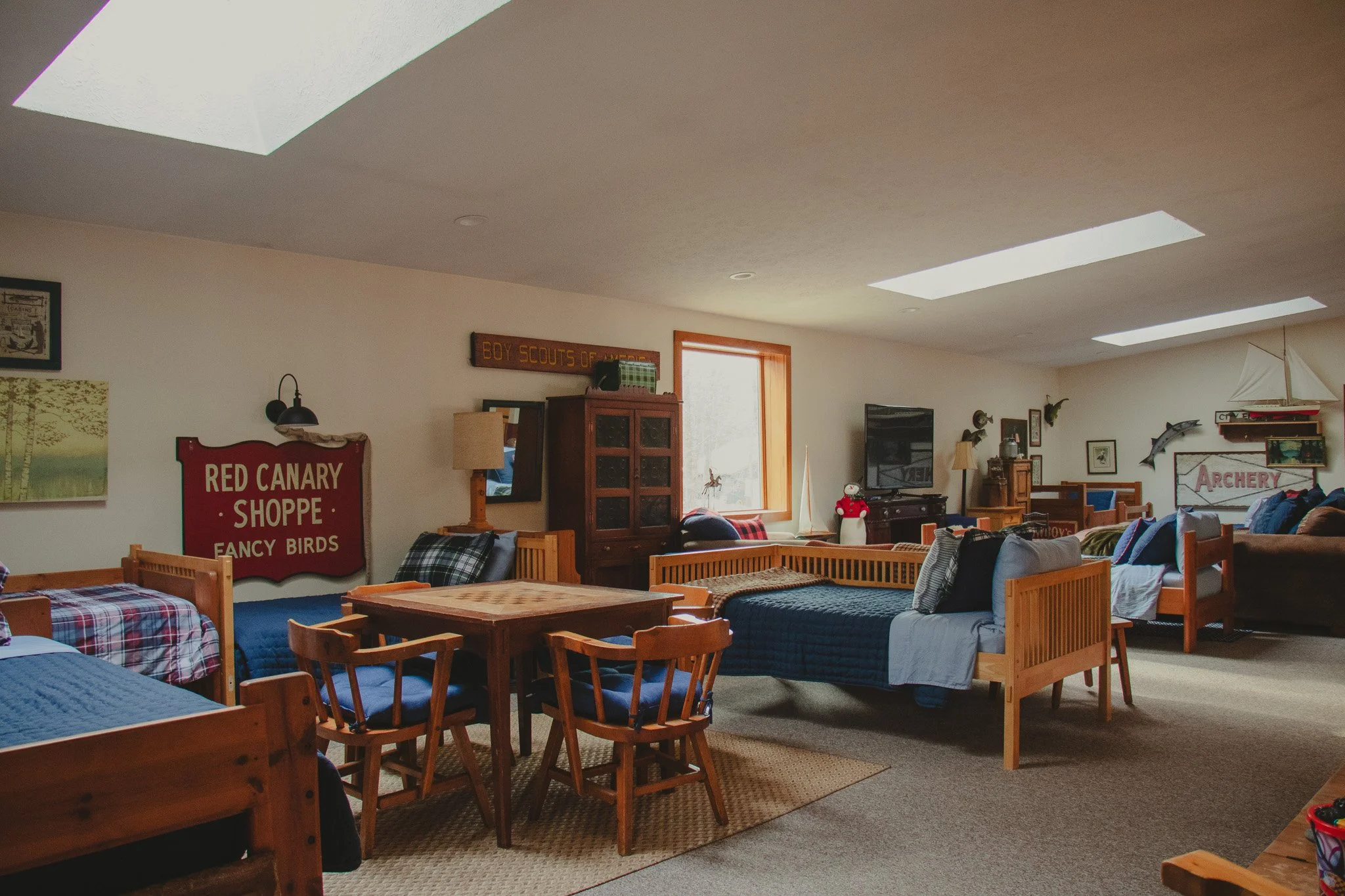 Indoor room with multiple beds, wooden furniture, framed pictures, and wall signs, including sign reading 'Red Canary Shoppe' and 'Boy Scouts of America