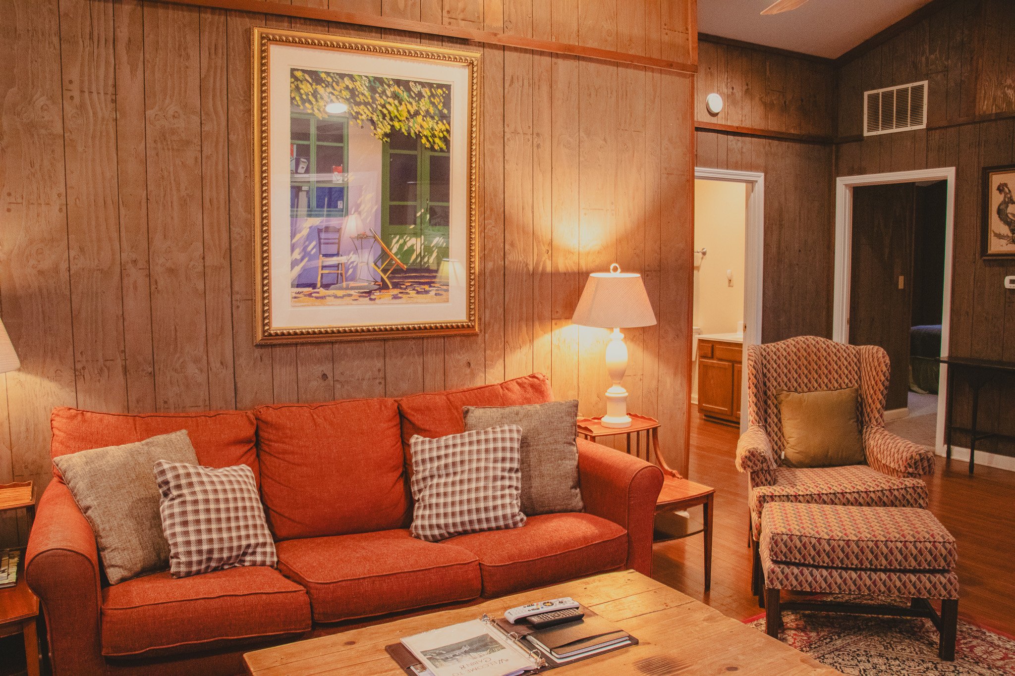 Living room with wood-paneled walls, featuring an orange sofa with multiple throw pillows, a wooden coffee table with magazines and a remote, a side table with a lamp, an armchair with a matching ottoman, and framed artwork on the walls.