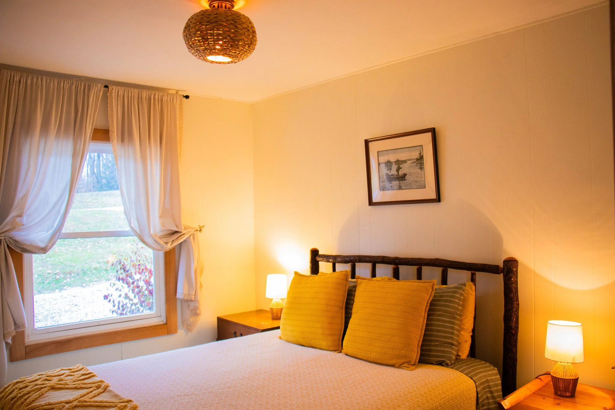 A cozy bedroom with a large bed with yellow and striped pillows, wooden nightstands with lamps, a window with white curtains, and a framed boat painting on the wall.