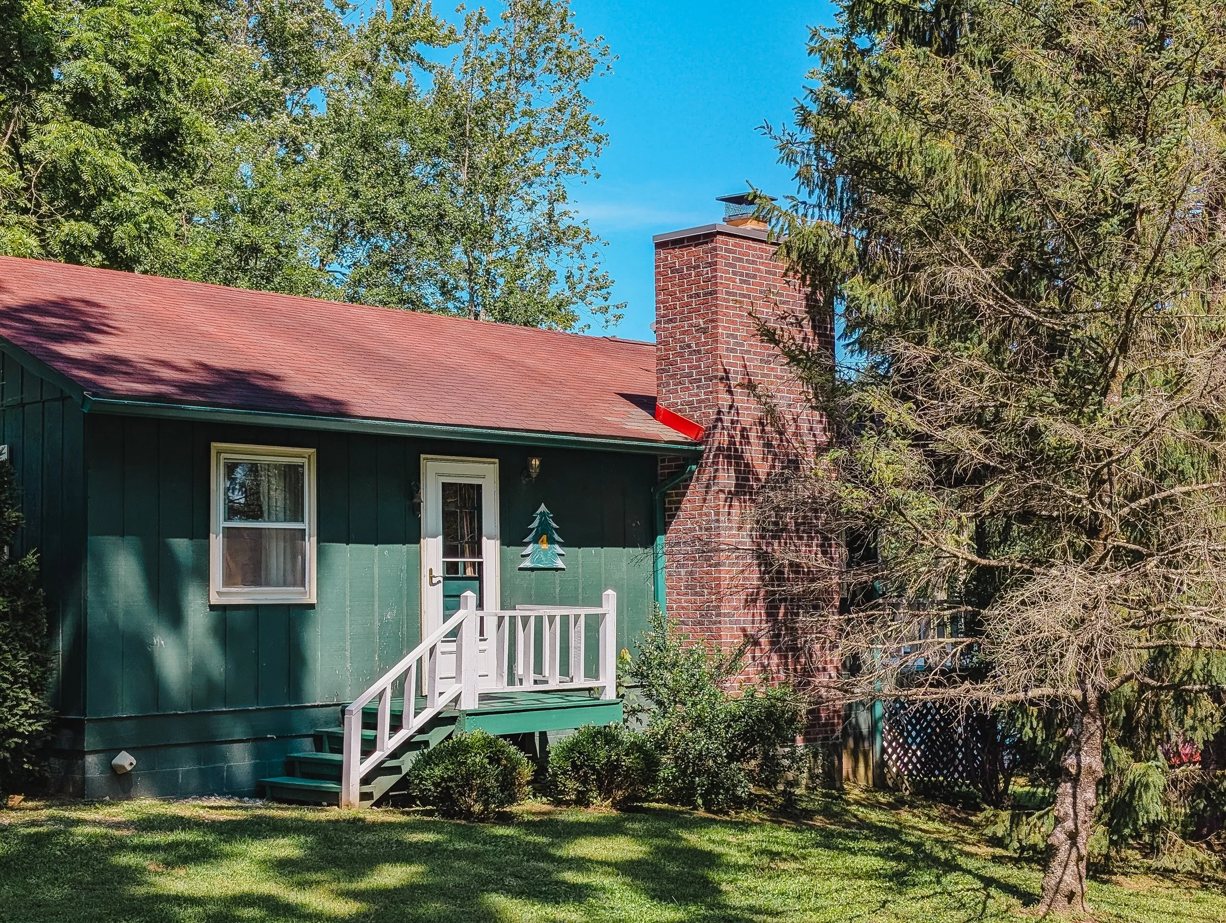 A small green house with a red shingled roof, a brick chimney, a small front porch with white railings, surrounded by greenery including trees and bushes, with a sunny blue sky in the background.