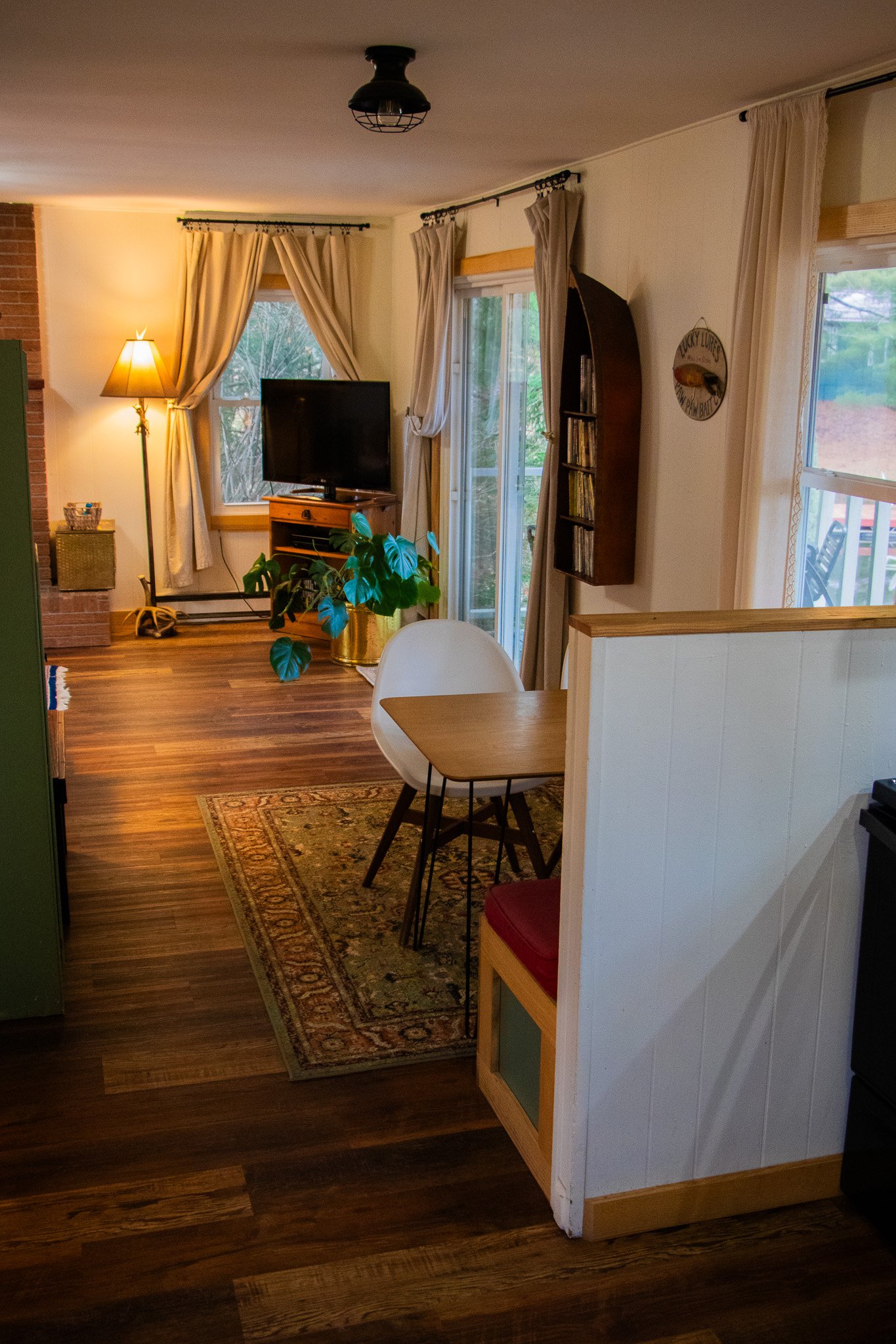 A cozy living room with hardwood floors, a window with curtains, a small dining table with a white chair, a TV on a wooden stand, a tall standing lamp, a potted plant, and a sliding glass door leading outside.