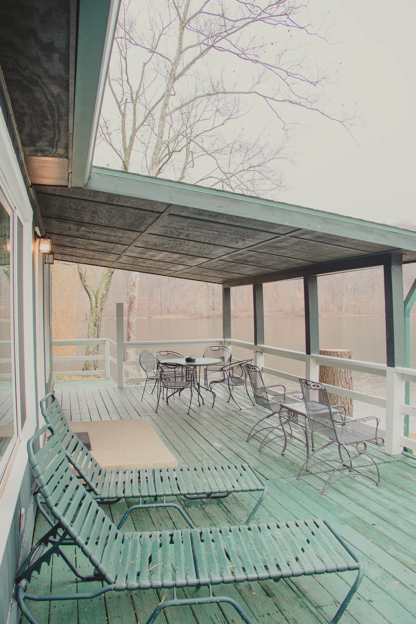 A covered outdoor deck overlooking a body of water with a leafless tree in the background. The deck has green wooden planks, two green lounge chairs, a round table with four chairs, and three additional chairs laid back on the deck.