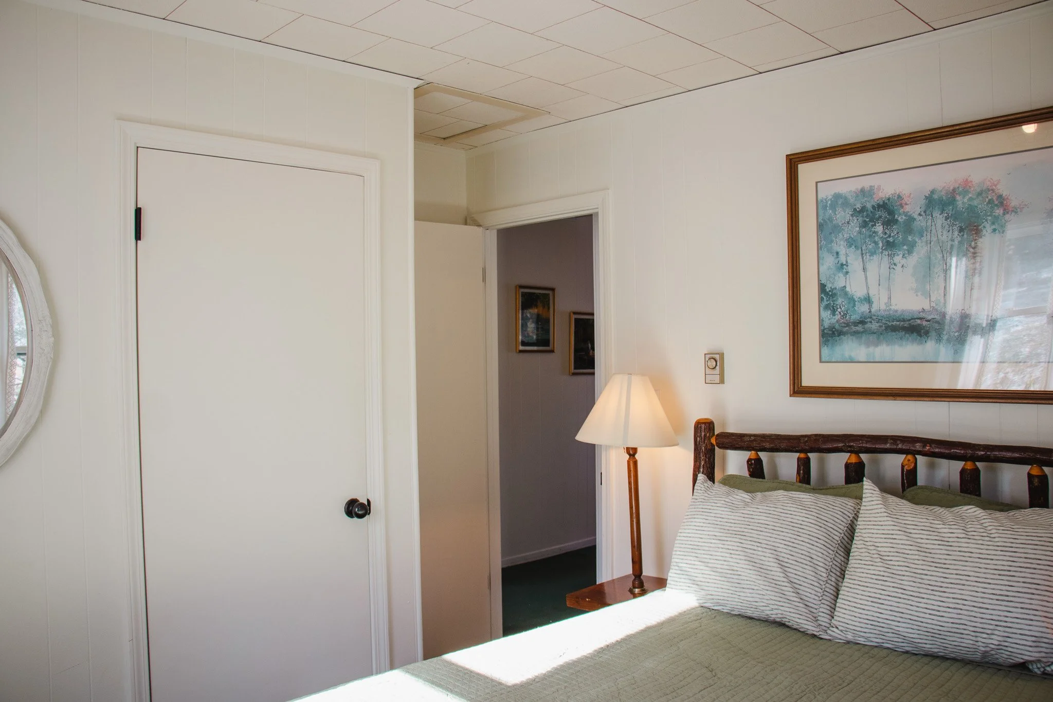 A cozy bedroom with a wooden bedframe, striped pillows, a table lamp, wall art, and a partially open door leading to a hallway with framed pictures.