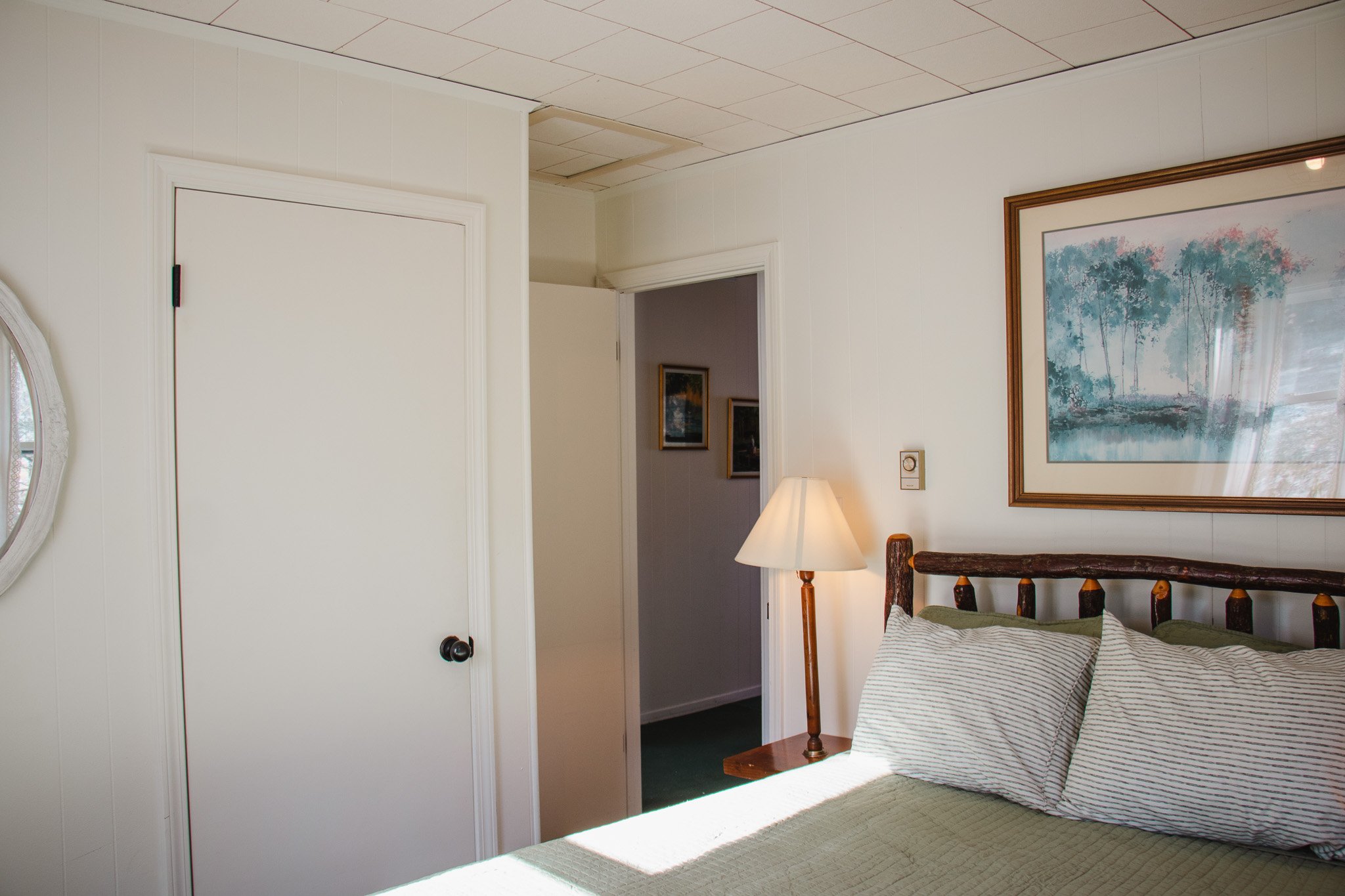 A cozy bedroom with a wooden bedframe, striped pillows, a table lamp, wall art, and a partially open door leading to a hallway with framed pictures.