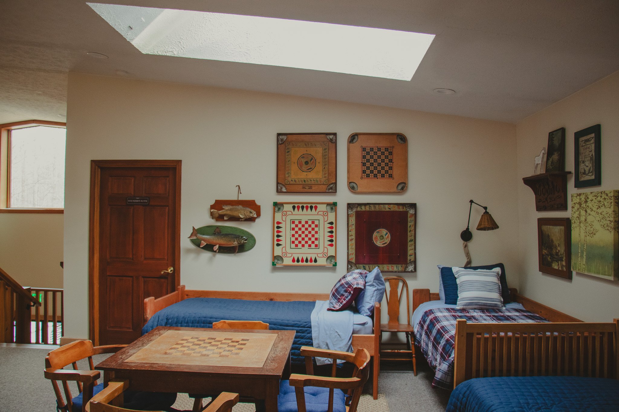 A cozy room with three beds, decorated with plaid and solid blue bedding. The wall features a collection of framed artwork and a mounted fish, emphasizing a rustic or cabin style. There's a wooden table and chairs in the foreground, and natural light