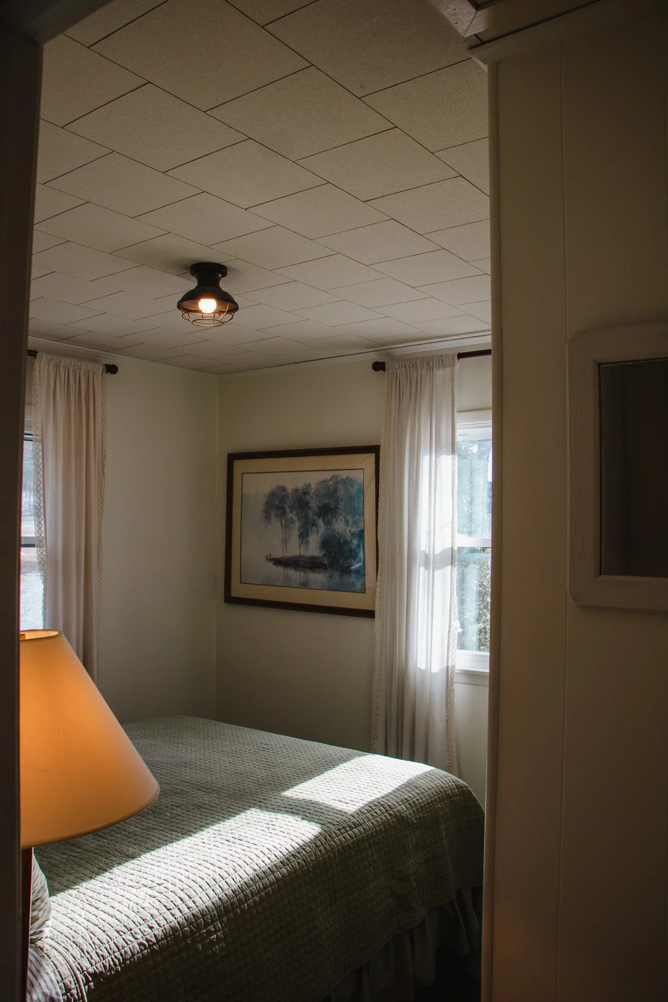 A cozy bedroom with a bed, a lamp, a framed painting of trees, and two windows with white curtains, sunlight streaming in.