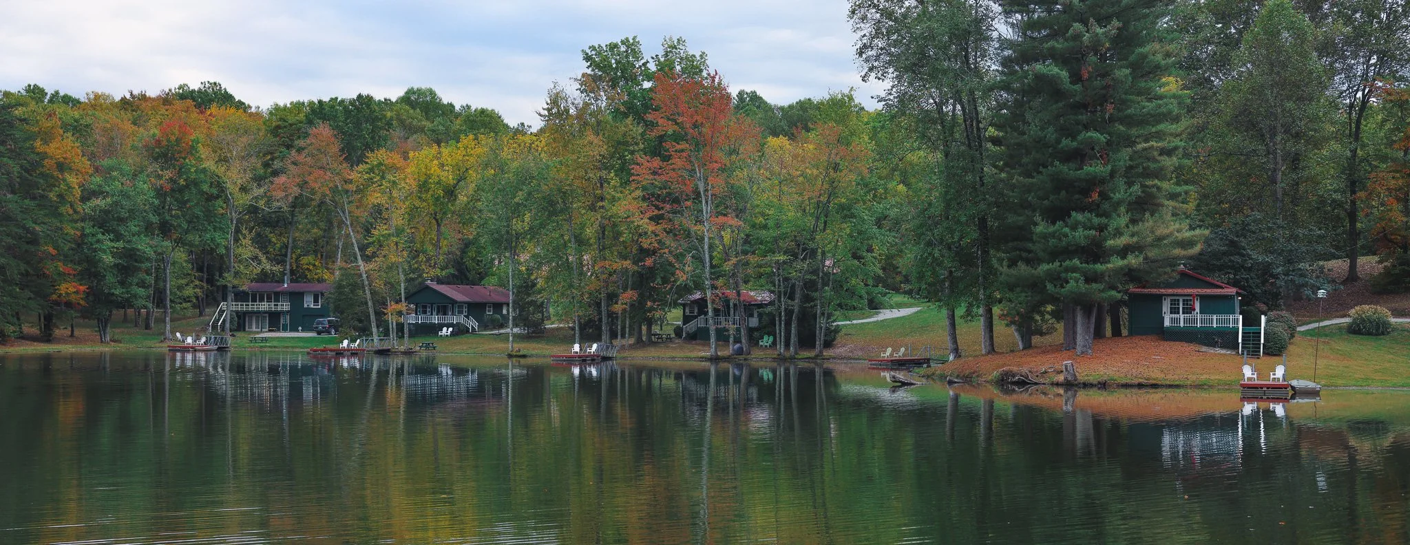 A peaceful lakeside scene with several small houses among trees displaying fall colors, some with red roofs, and docks with boats along the shore. The water reflects the trees and houses, and the sky is cloudy.