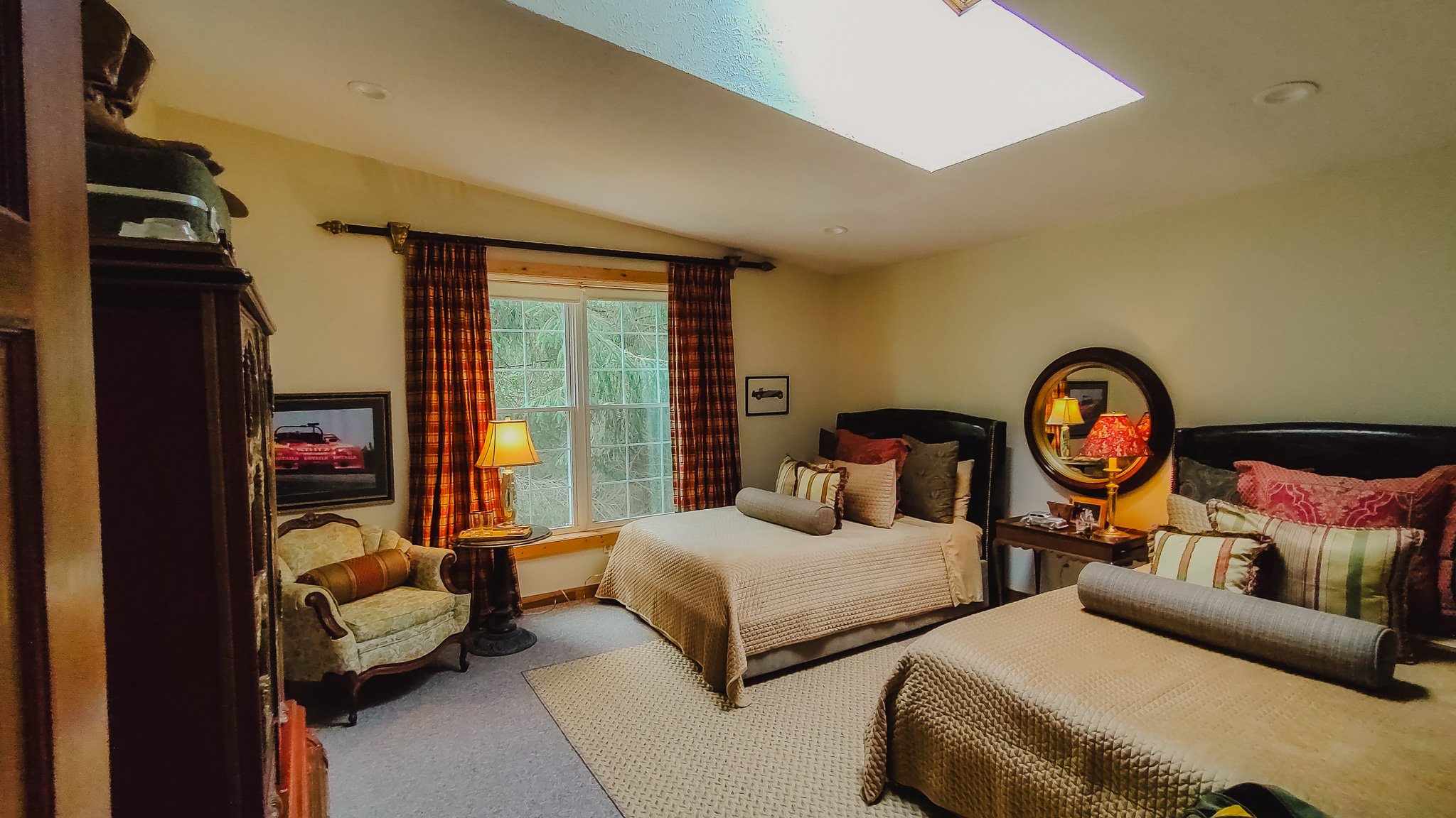 A cozy bedroom with two beds, each with multiple pillows, a window with plaid curtains, a round mirror above the nightstand, a lamp, a framed picture of a car, and a vintage loveseat.
