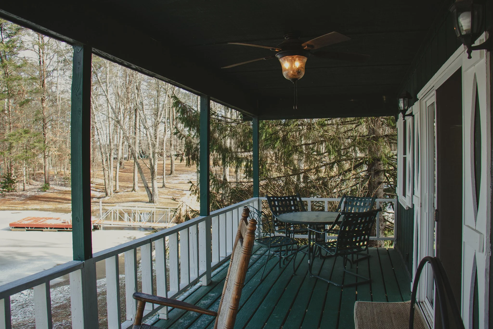 Covered porch overlooking trees and partly frozen pond, with outdoor furniture including a round table and chairs.