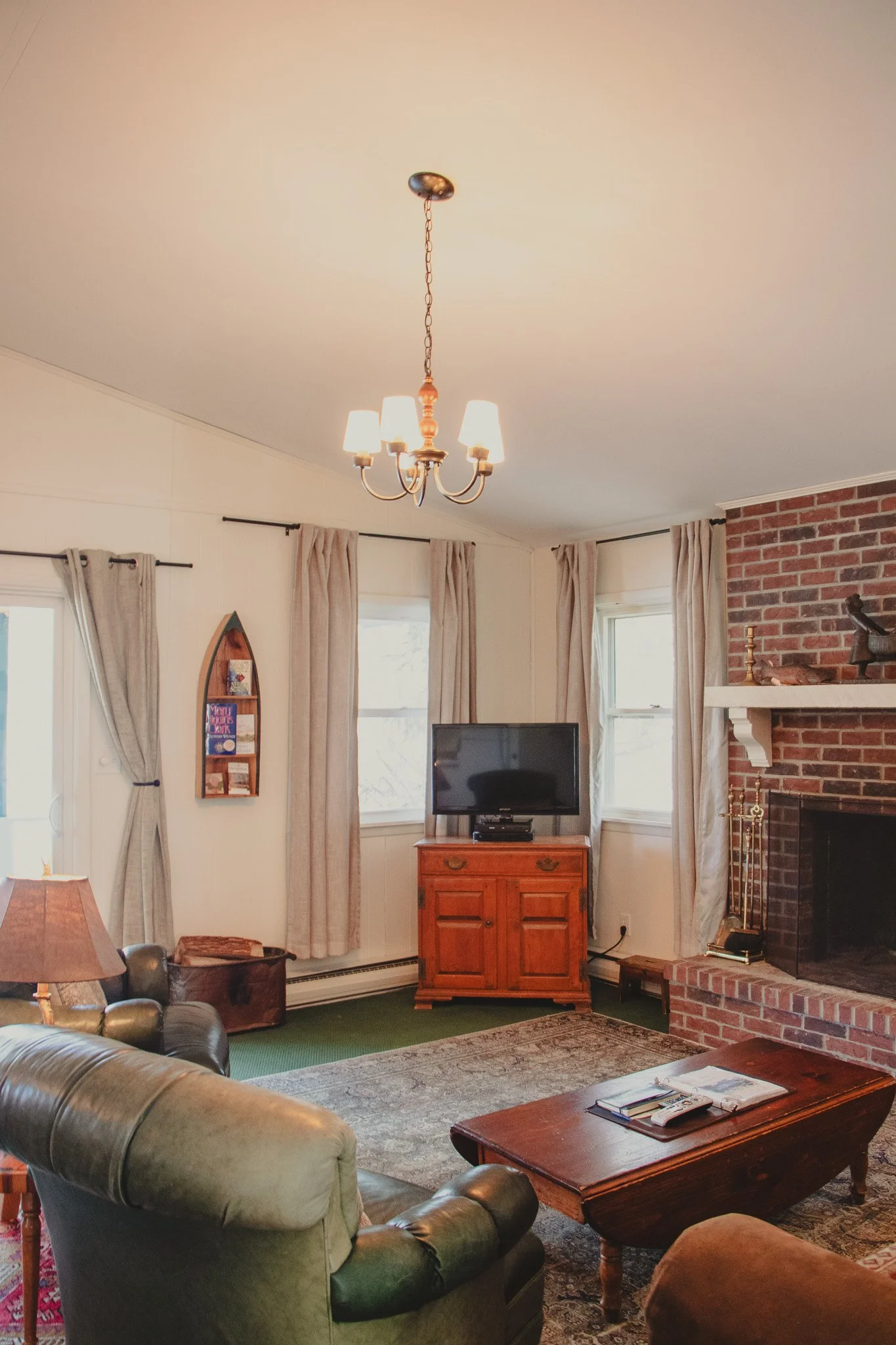 Living room with a brick fireplace, wooden furniture, sofa, armchair, coffee table, TV, and windows with curtains.