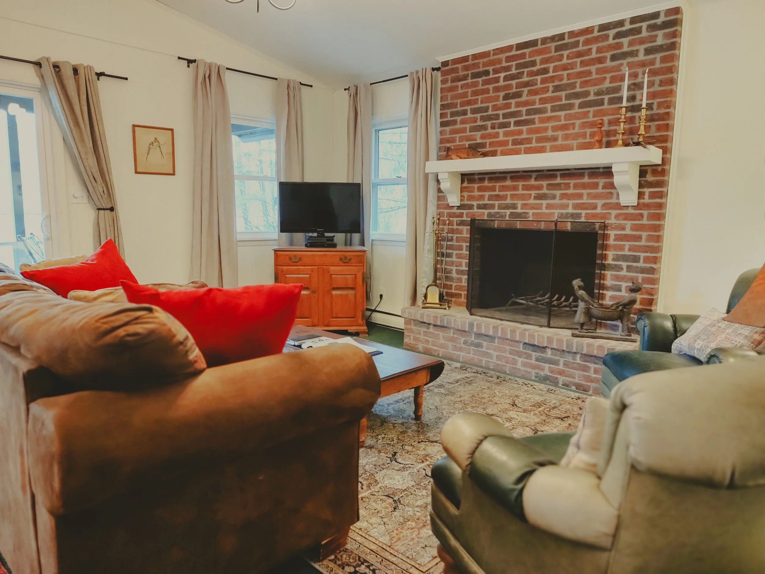 Living room with a brick fireplace, green and brown upholstered chairs, a wooden coffee table, a red cushion, a wooden TV stand, a small flat-screen TV, and windows with beige curtains.