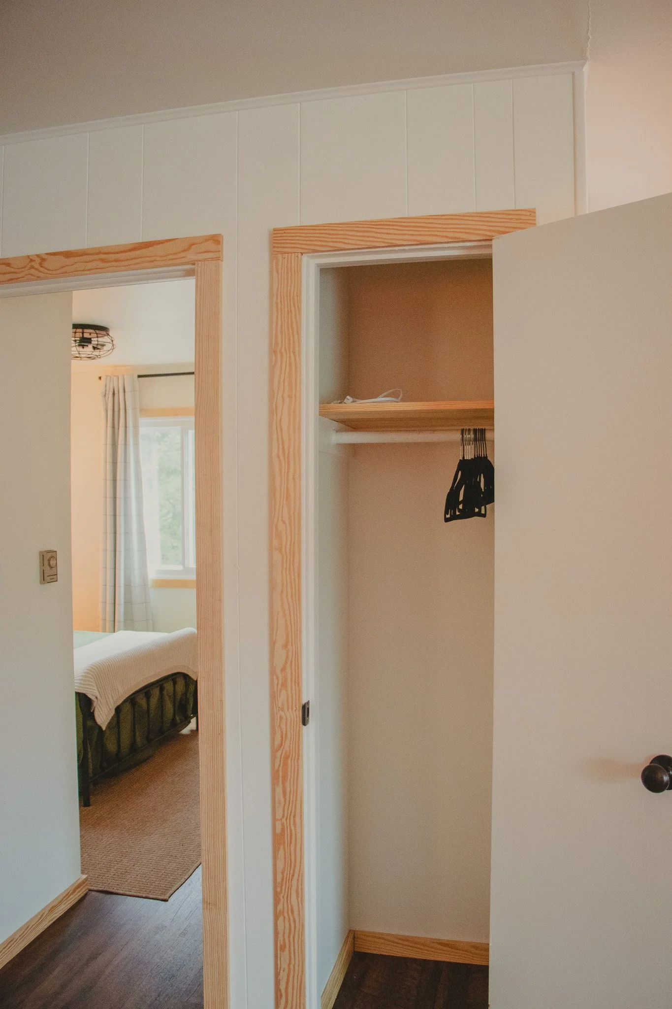 Empty closet with a wooden shelf and a few black hangers, next to a bedroom with white curtains and a bed.