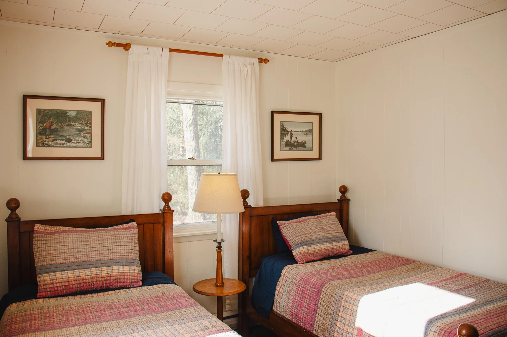 A bedroom with two twin beds featuring wooden headboards, plaid bedspreads, and matching pillows. A wooden nightstand with a lamp is placed between the beds. There is a window with white curtains and two framed pictures of outdoor scenes hanging on t