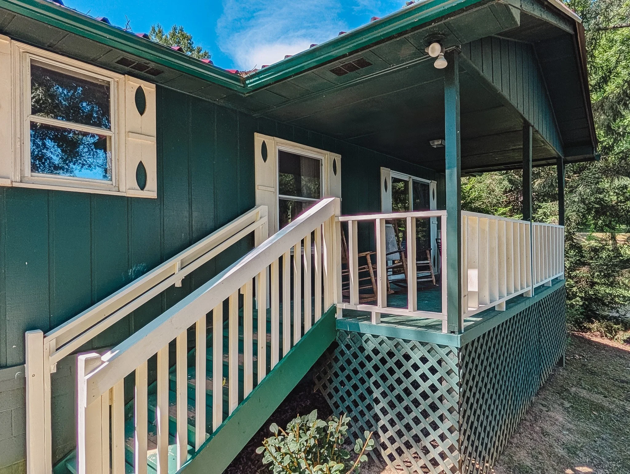 Front porch of a dark green house with white trim, a white railing, and lattice underneath, featuring three rocking chairs and stairs leading up from the ground.