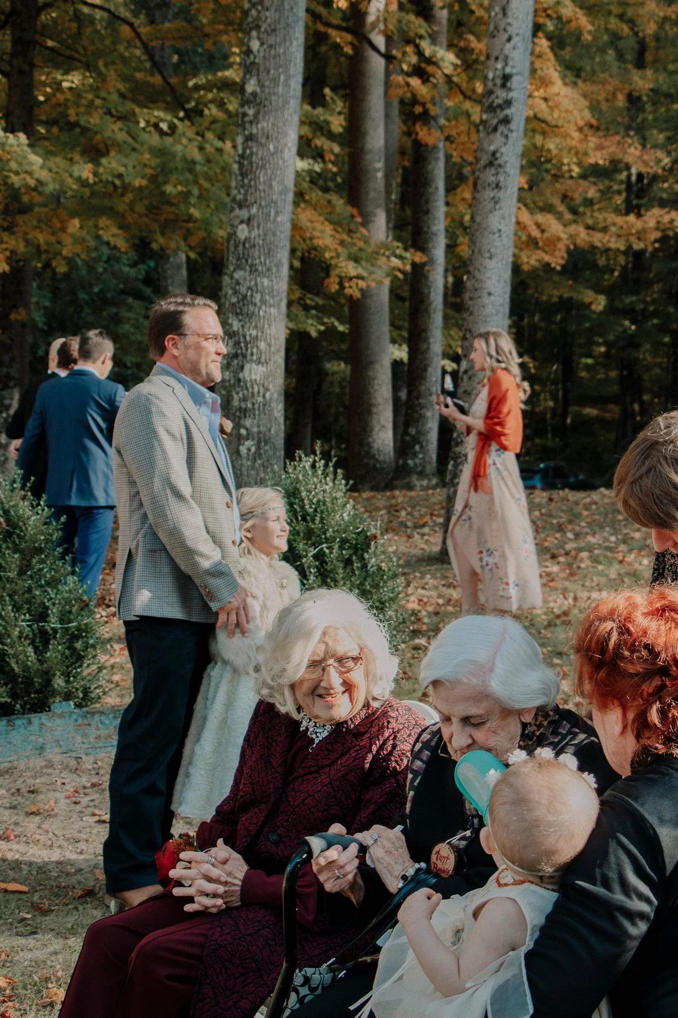 A group of people outdoors during fall, with children and elderly women, some talking and others using their phones, in a wooded area with orange and green leaves.