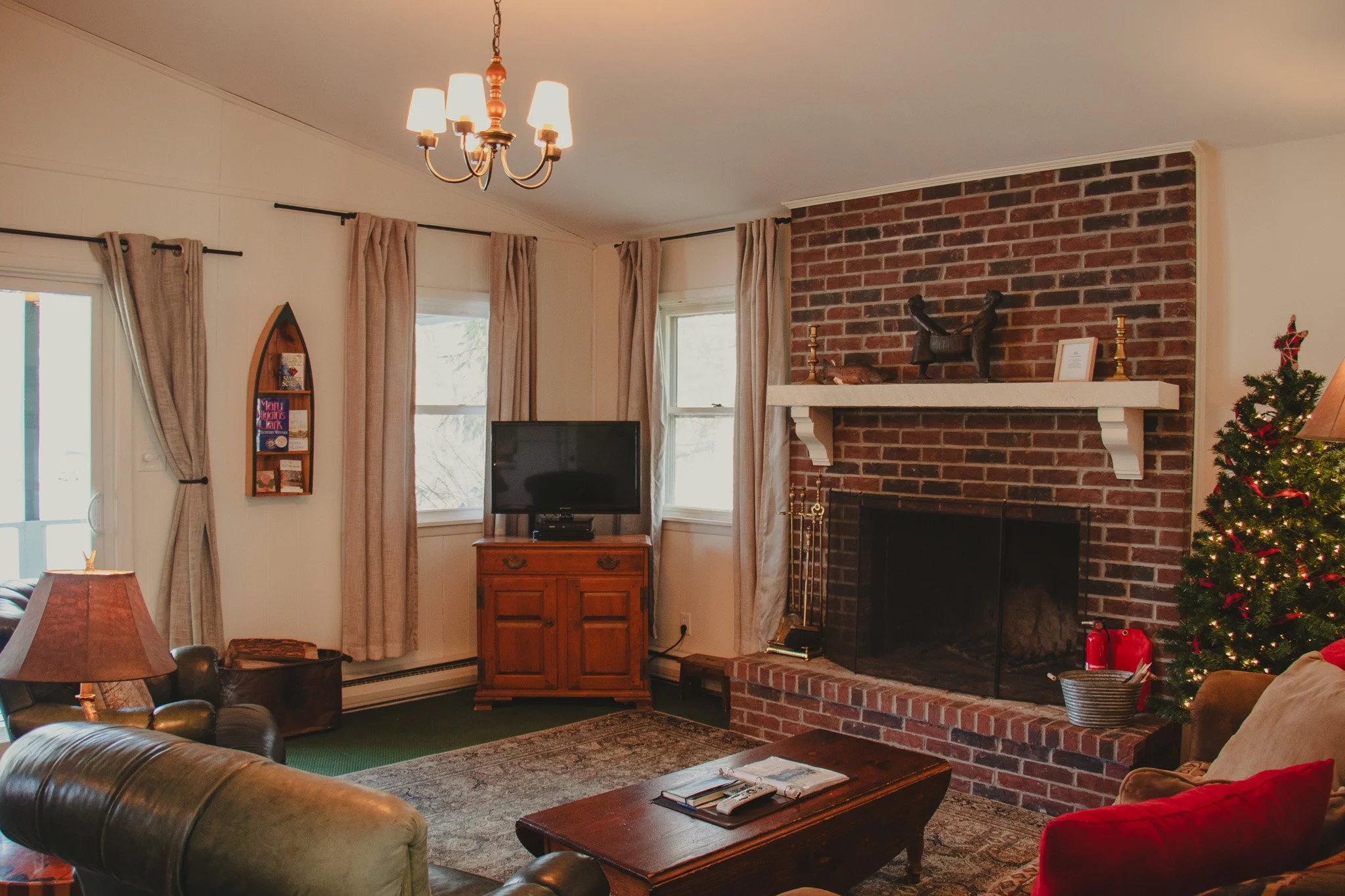 Living room with a brick fireplace, a decorated Christmas tree, a sofa with red and tan cushions, a wooden coffee table, a flat-screen TV on a wooden stand, a window with beige curtains, a sliding glass door, and a chandelier.