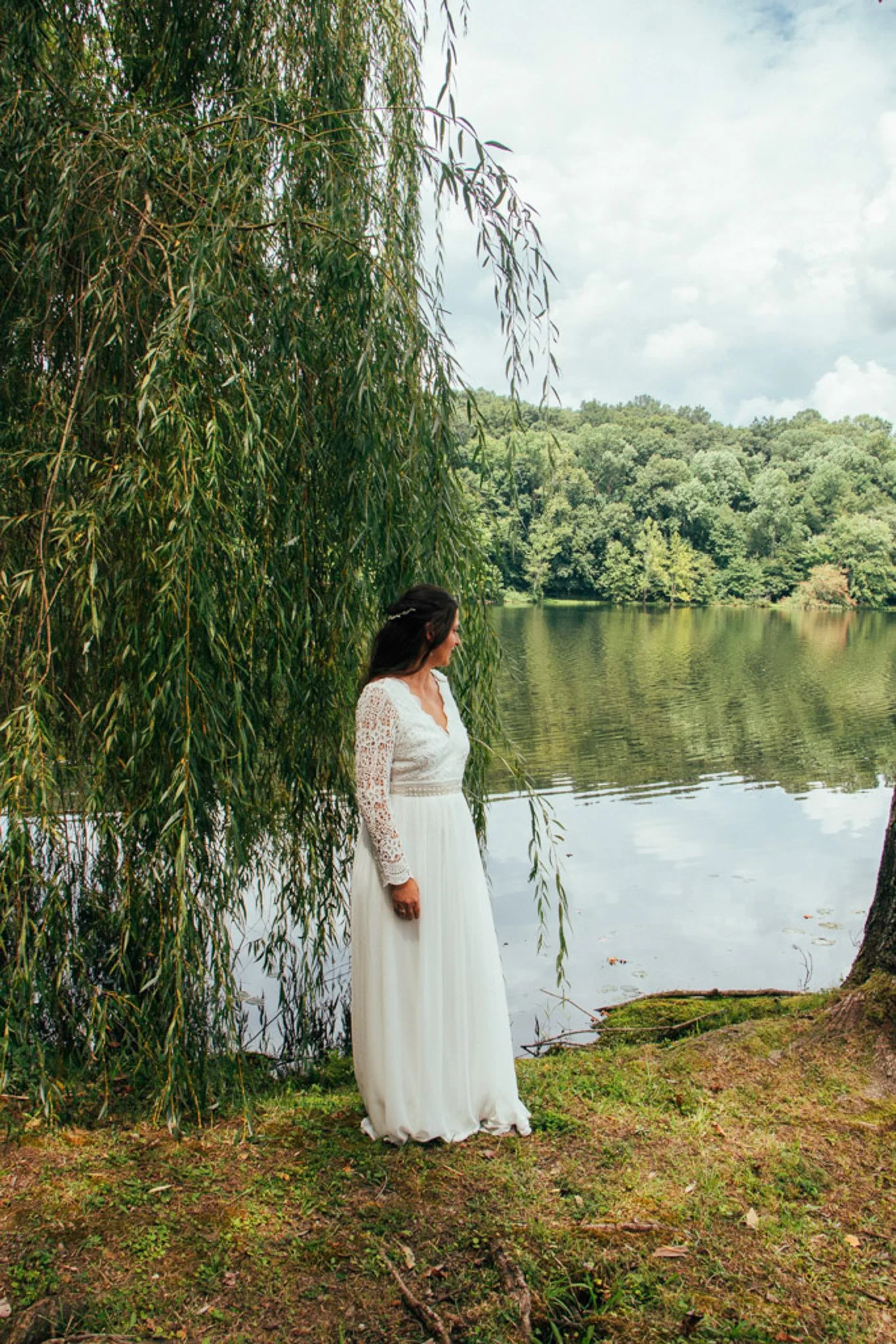 A woman in a white dress with lace sleeves standing by a lake, surrounded by lush green trees with a weeping willow hanging down over the water.
