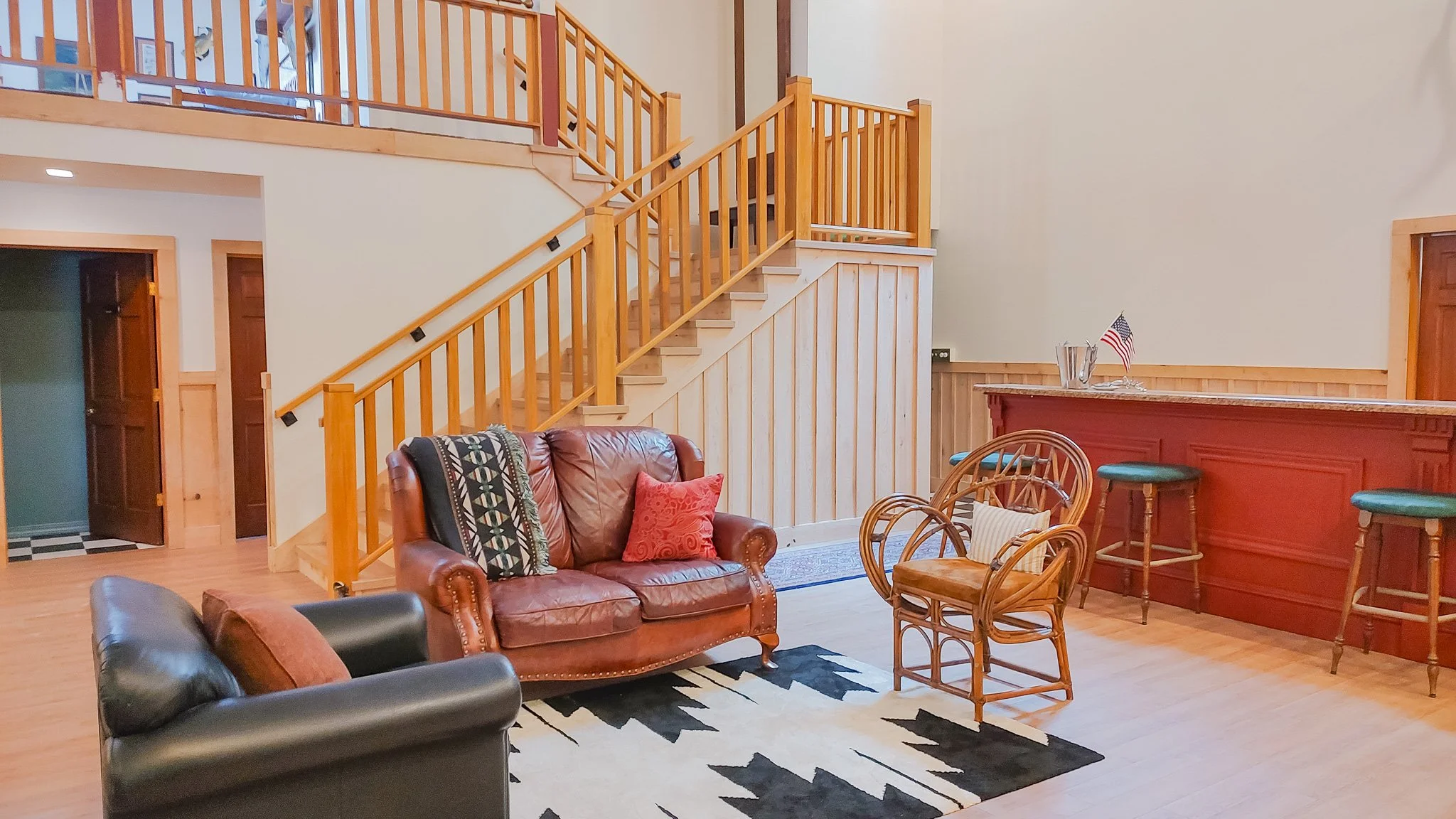 Living room with leather sofa and armchairs, wooden staircase with railing, and a bar area with stools, American flag, and decorative items.