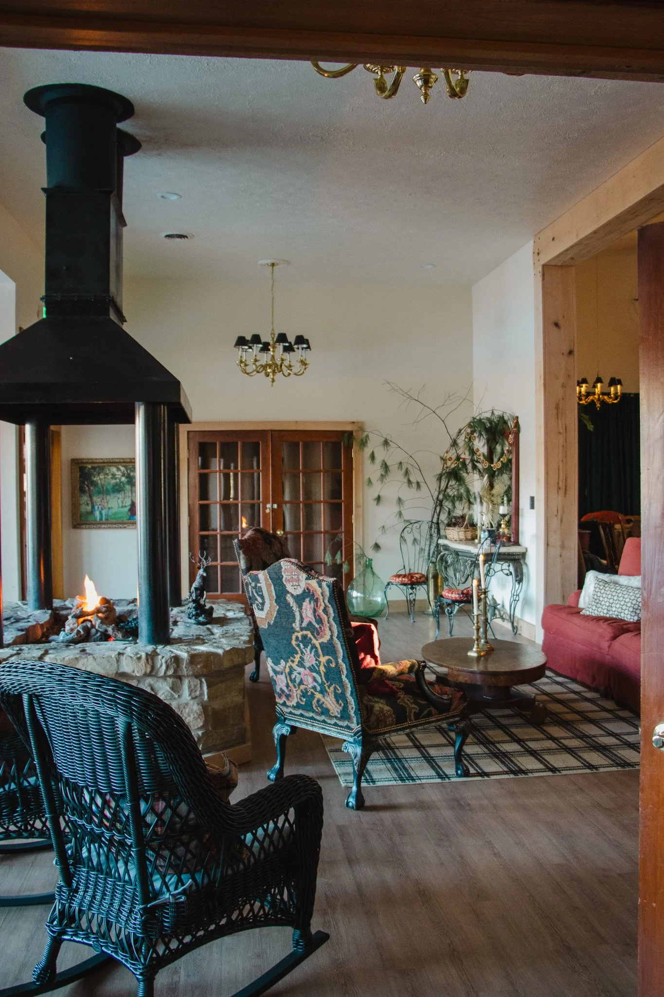 Cozy living room with a stone fireplace, wicker rocking chair, upholstered armchair, wooden coffee table, pink sofa, vintage dresser, and decorative plants.