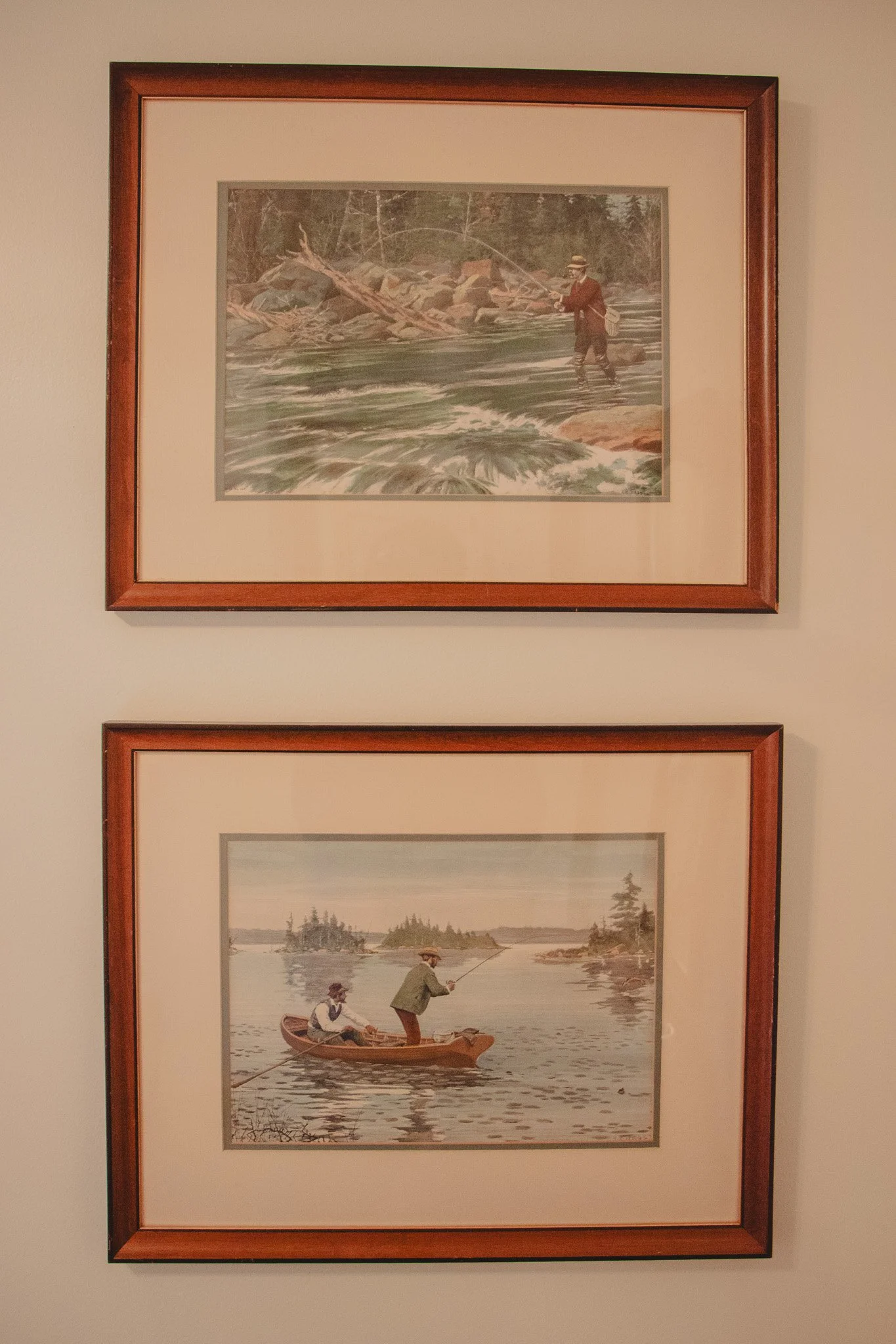 Two framed watercolor paintings of people fishing on a river, mounted on a wall. The top painting depicts a man standing in the water fishing, and the bottom painting shows a man and a woman fishing from a boat.