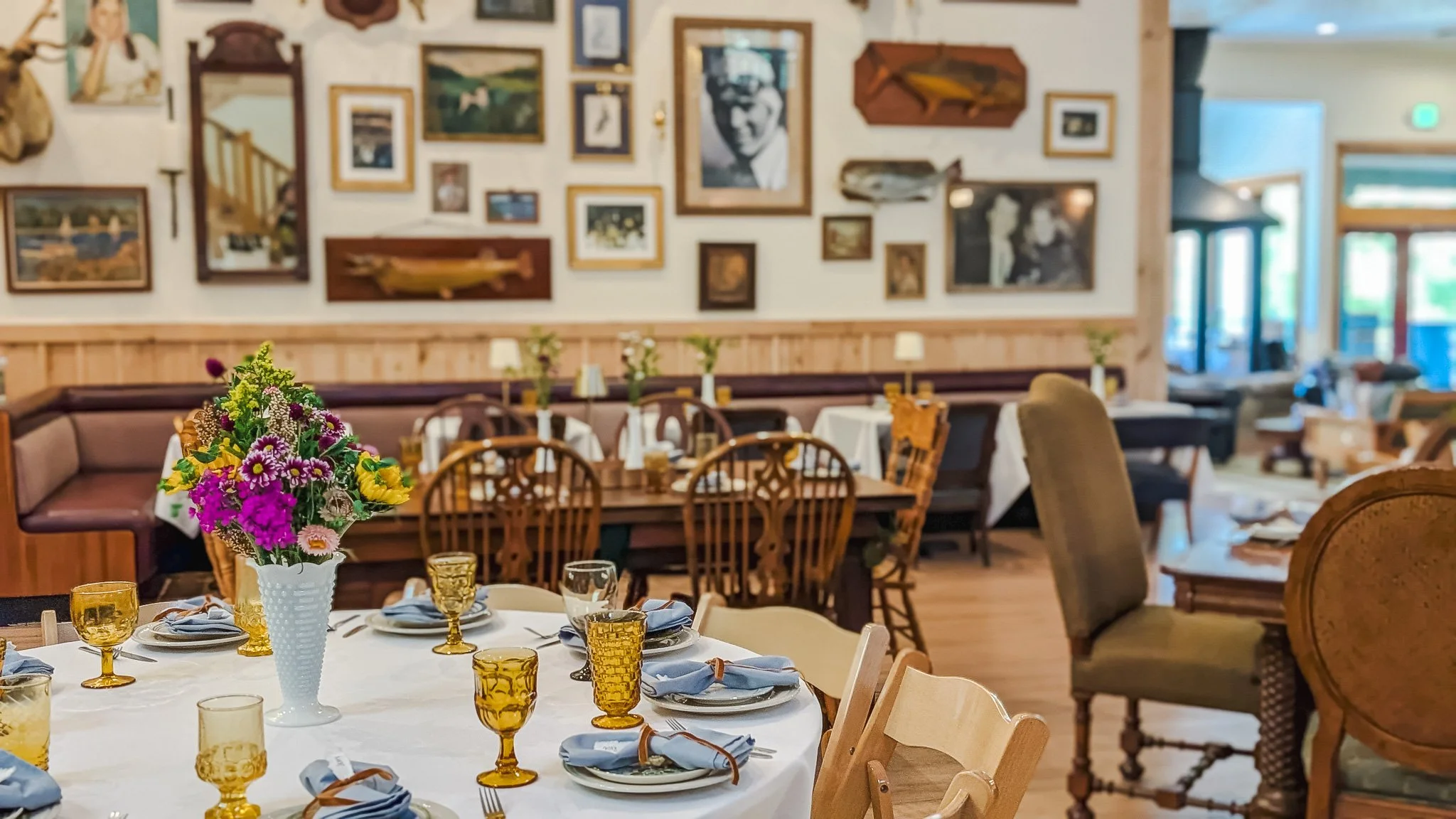 A dining area with a round table set with yellow glasses, blue napkins, and a vase of colorful flowers. The background features a wall decorated with various framed pictures and a mix of wooden chairs and upholstered seating.