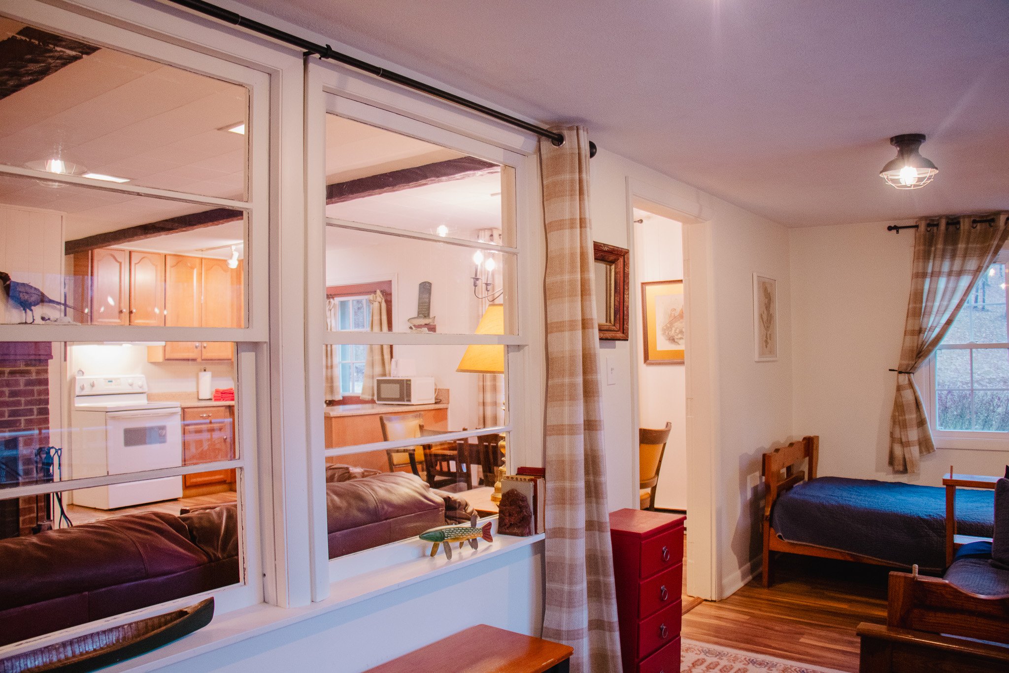 View of a living room with a window looking into a kitchen, a bedroom with a bed, a red drawer, and curtains.