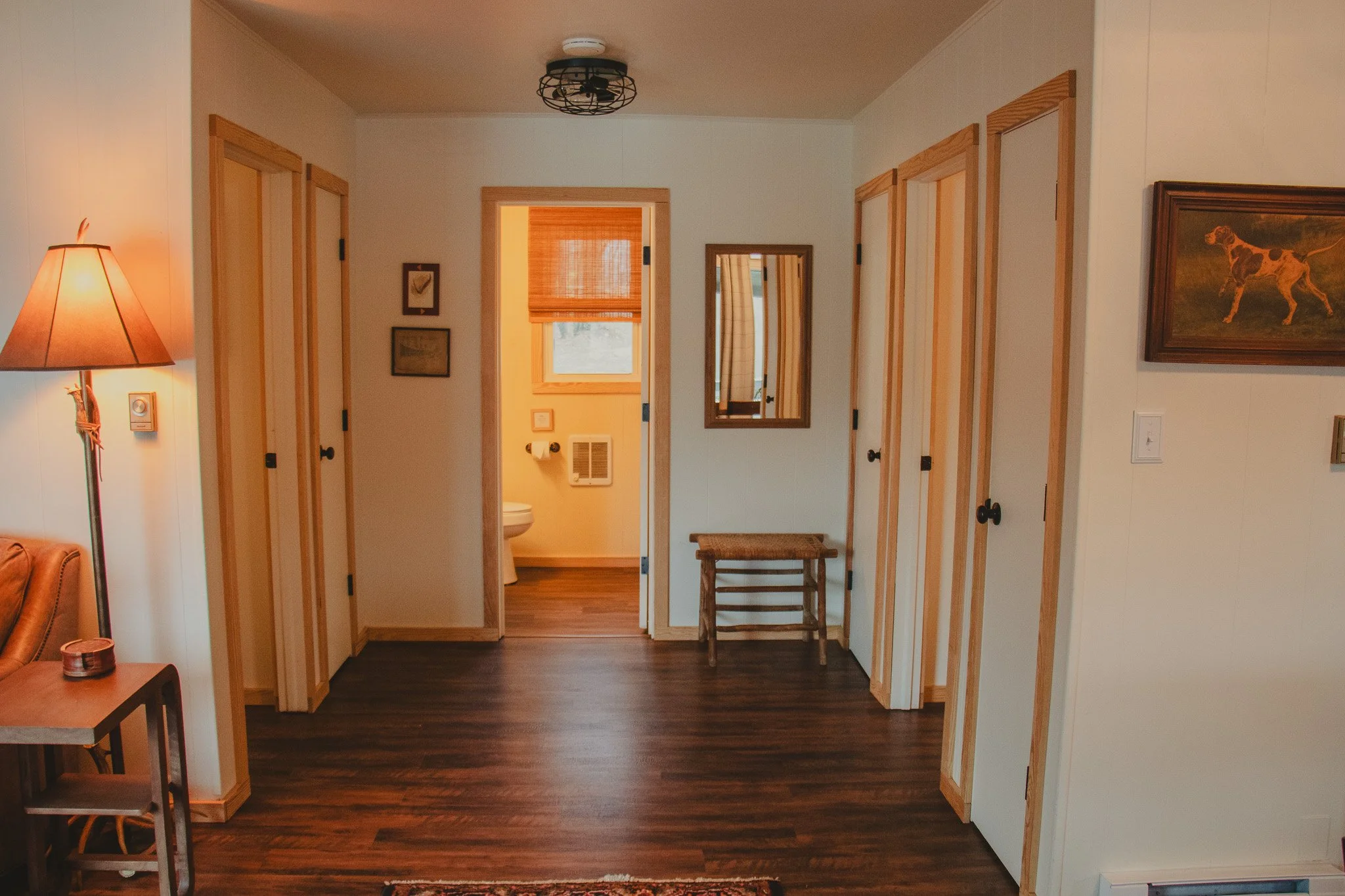 Interior view of a cozy room with wooden floors, a small wooden bench, and three closed doors on the right. A wall mirror is next to the bathroom, which is visible through an open door, with a window with a woven shade. The room features warm lightin