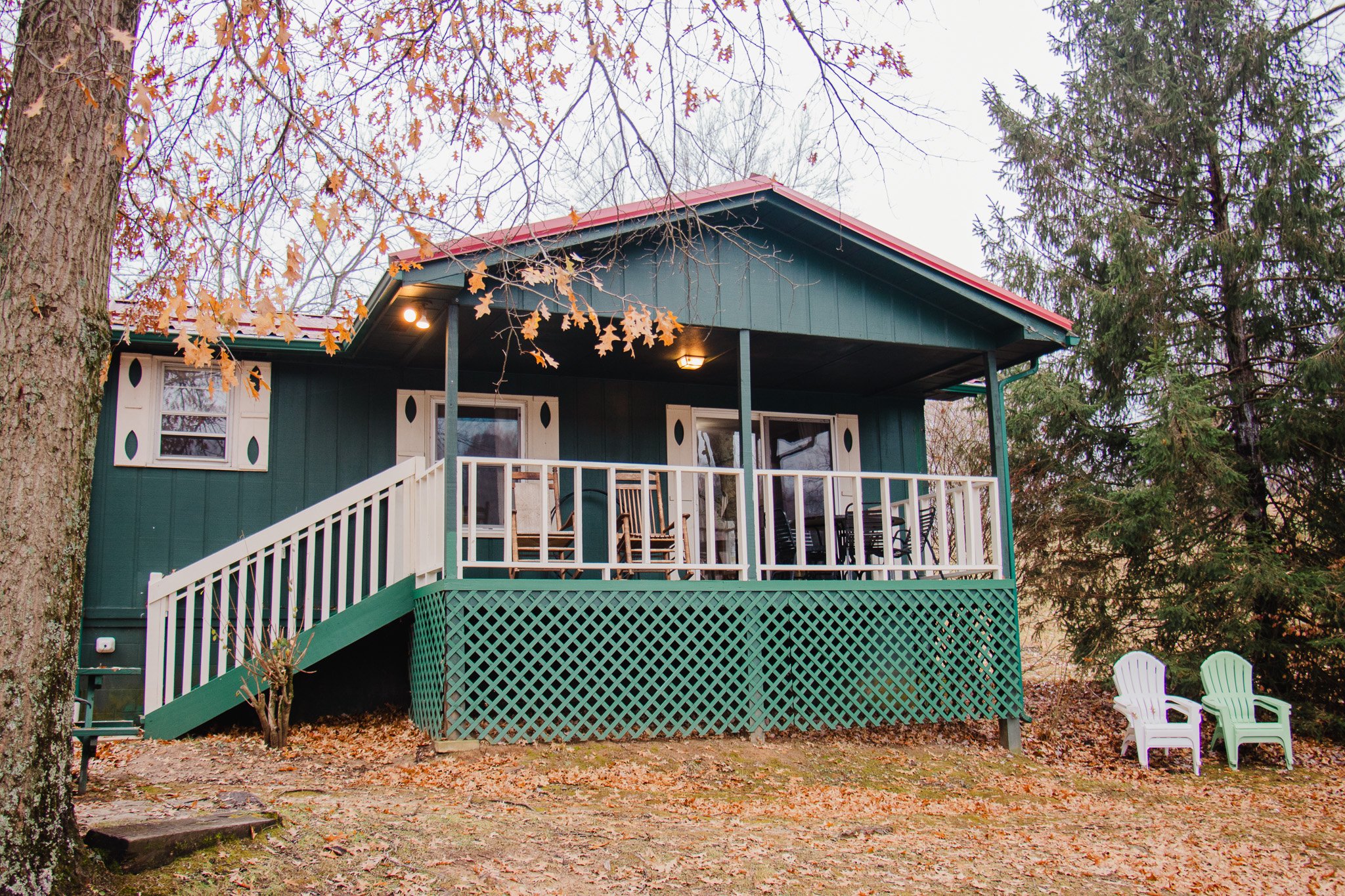A house with a green exterior and white balcony rails, surrounded by trees with autumn leaves, two white and one green Adirondack chairs on the ground outside.