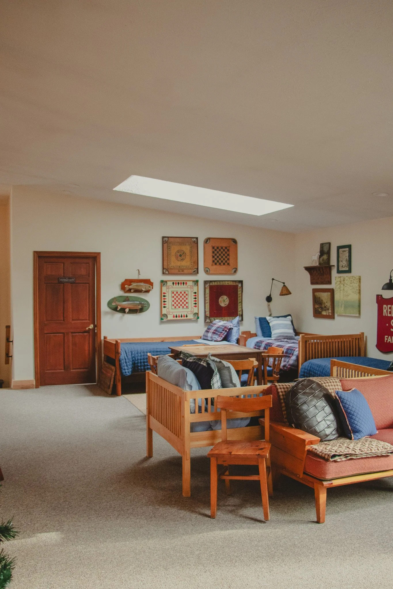 A cozy room with wooden furniture, twin beds with blue and plaid bedding, a pink sofa with pillows, and a wall decorated with vintage game boards and paintings, under a skylight.