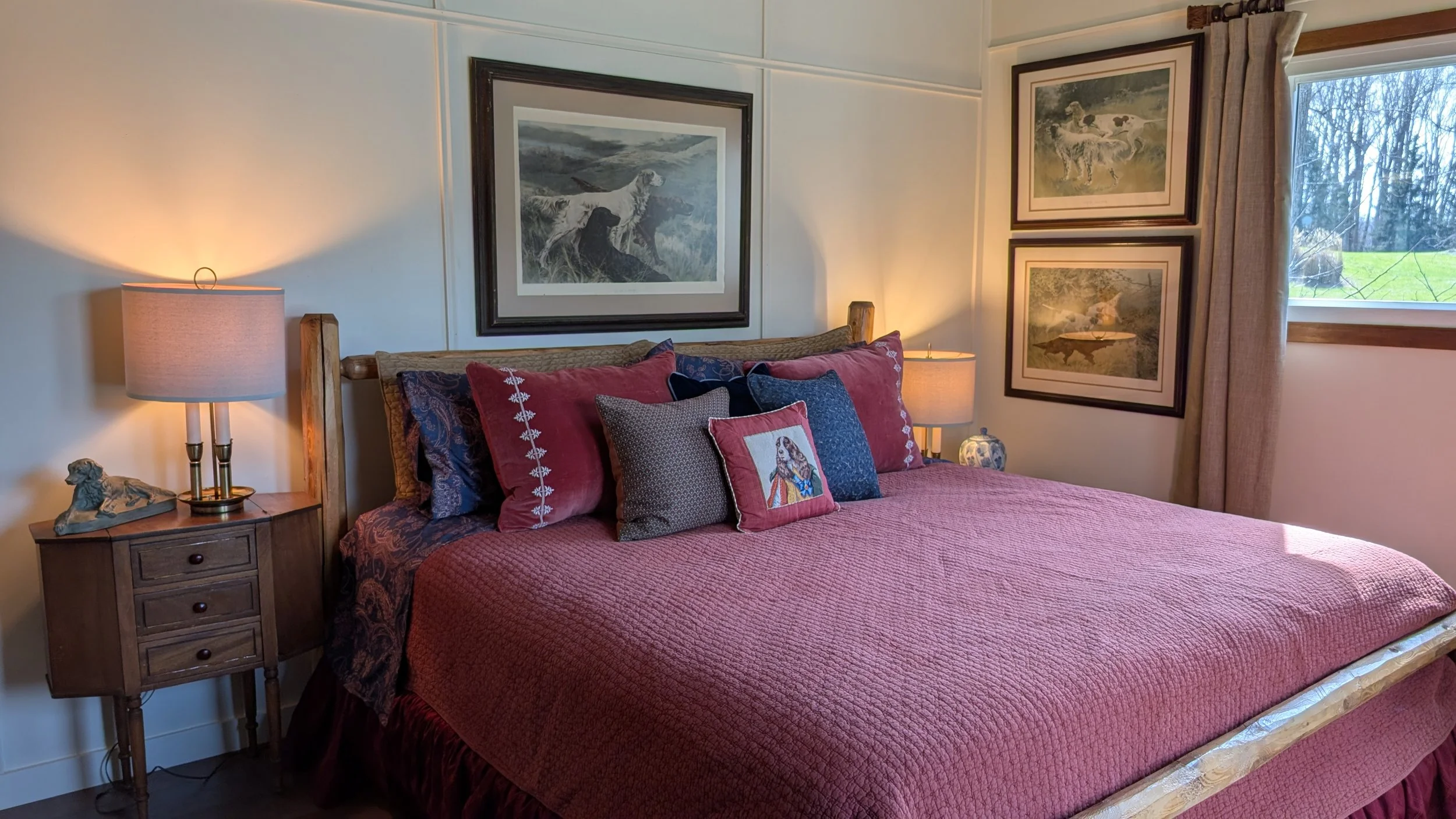 A cozy bedroom with wood-paneled walls, a king-size bed with rust-colored bedding and pillows, two bedside tables with lamps, framed hunting art prints on the wall, a window with curtains showing an outdoor scene with trees.