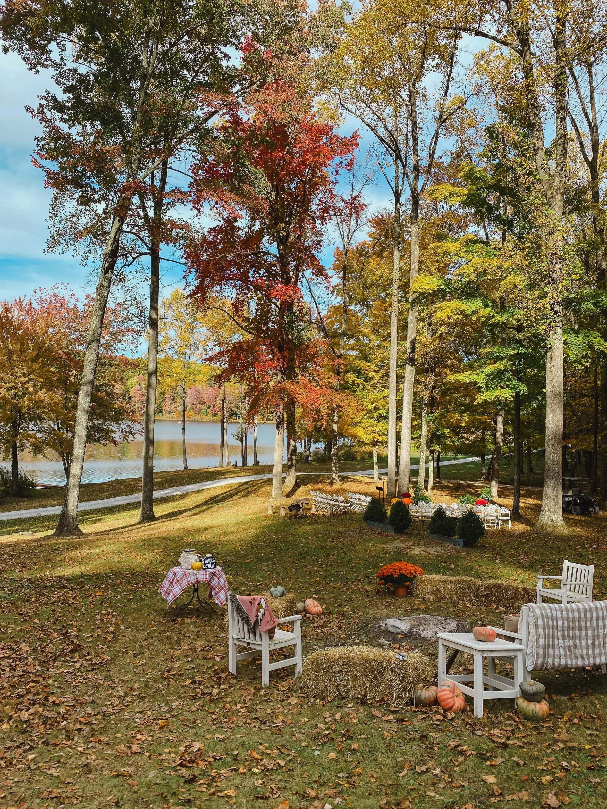Outdoor autumn scene with decorated chairs, pumpkins, trees with fall foliage, and a lake in the background.
